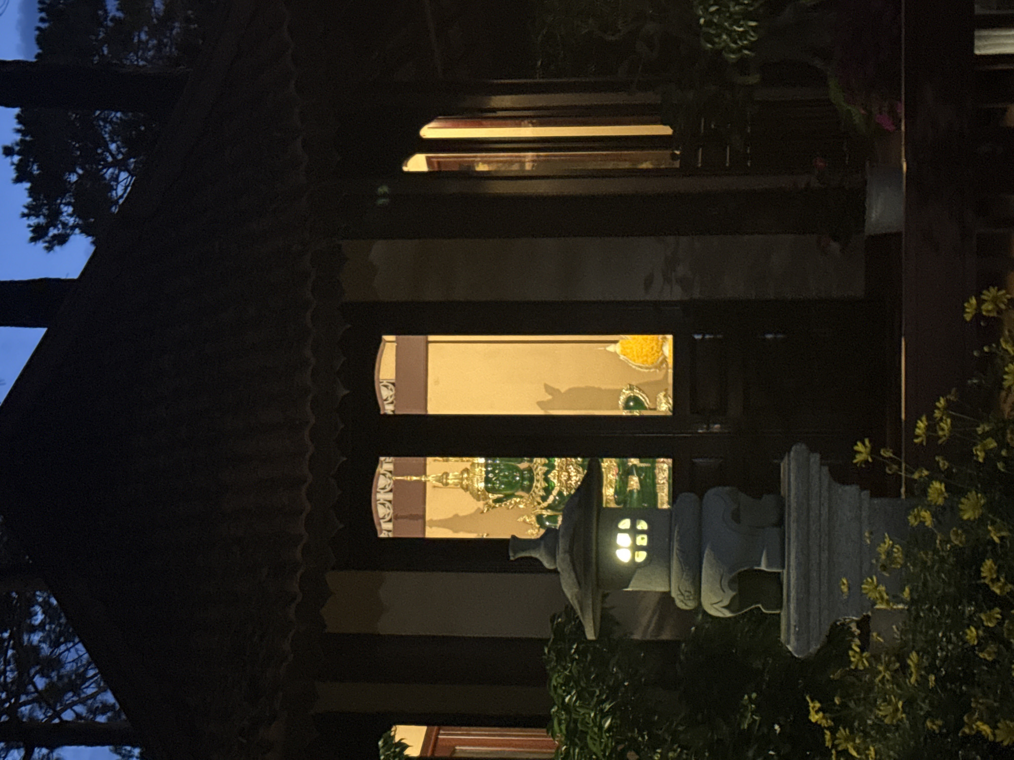 This image captures a nighttime exterior view of a building in Da Lat, Vietnam, displaying elements of traditional Asian architecture, likely a temple or pagoda. The building features a dark, multi-tiered roof with scalloped eaves, silhouetted against a dark blue evening sky. Dark, tall trees, possibly pines, are visible in silhouette against the sky in the upper background.
The central part of the building shows illuminated interiors through multiple windows and a central doorway. A prominent green statue, possibly crafted from jade or a similar material, with an elaborate golden headdress and adornments, is visible through the central opening. To its right, a smaller yellow conical object is present. The interior illumination is warm-toned.
In the lower-left foreground, a stone lantern (toro) with illuminated square openings stands on a stone pedestal, emitting a soft, warm light. Surrounding the base of the lantern and extending into the lower-left are clusters of small yellow flowers. Dark green foliage and bushes are visible around the base of the building and behind the lantern. To the right, a dark wooden railing or platform is present, with a white potted plant and additional dark foliage. No people are visible in the scene.