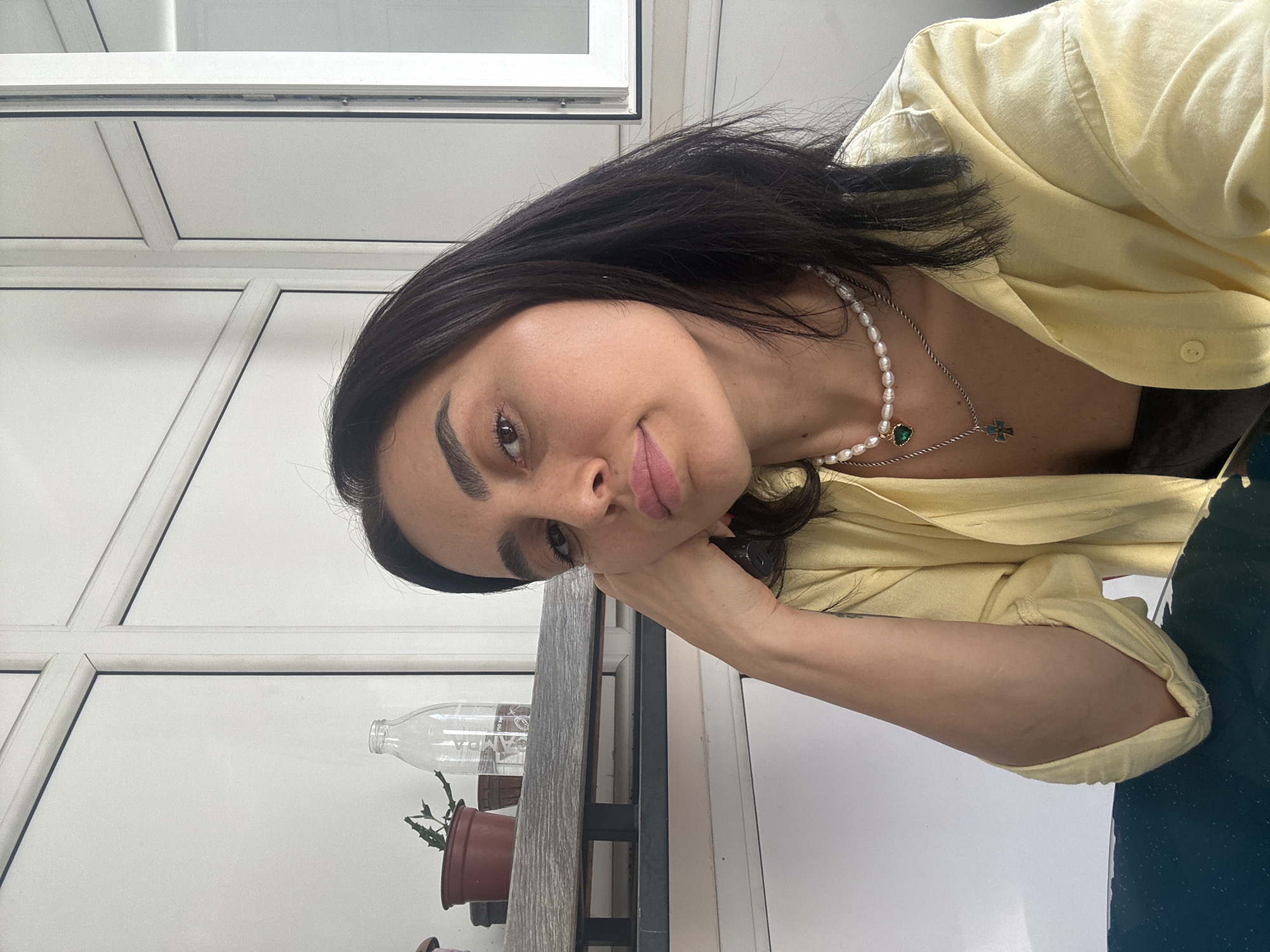 A medium shot captures a woman resting her head on her hand, looking at the camera. She has dark hair, dark eyebrows, and is wearing a light yellow shirt. A pearl necklace with a green pendant and a silver chain with a cross pendant adorn her neck. The background appears to be an indoor space with white walls and a window. A shelf with plants and a glass bottle is visible behind her. The overall lighting suggests daytime. The location is unknown.