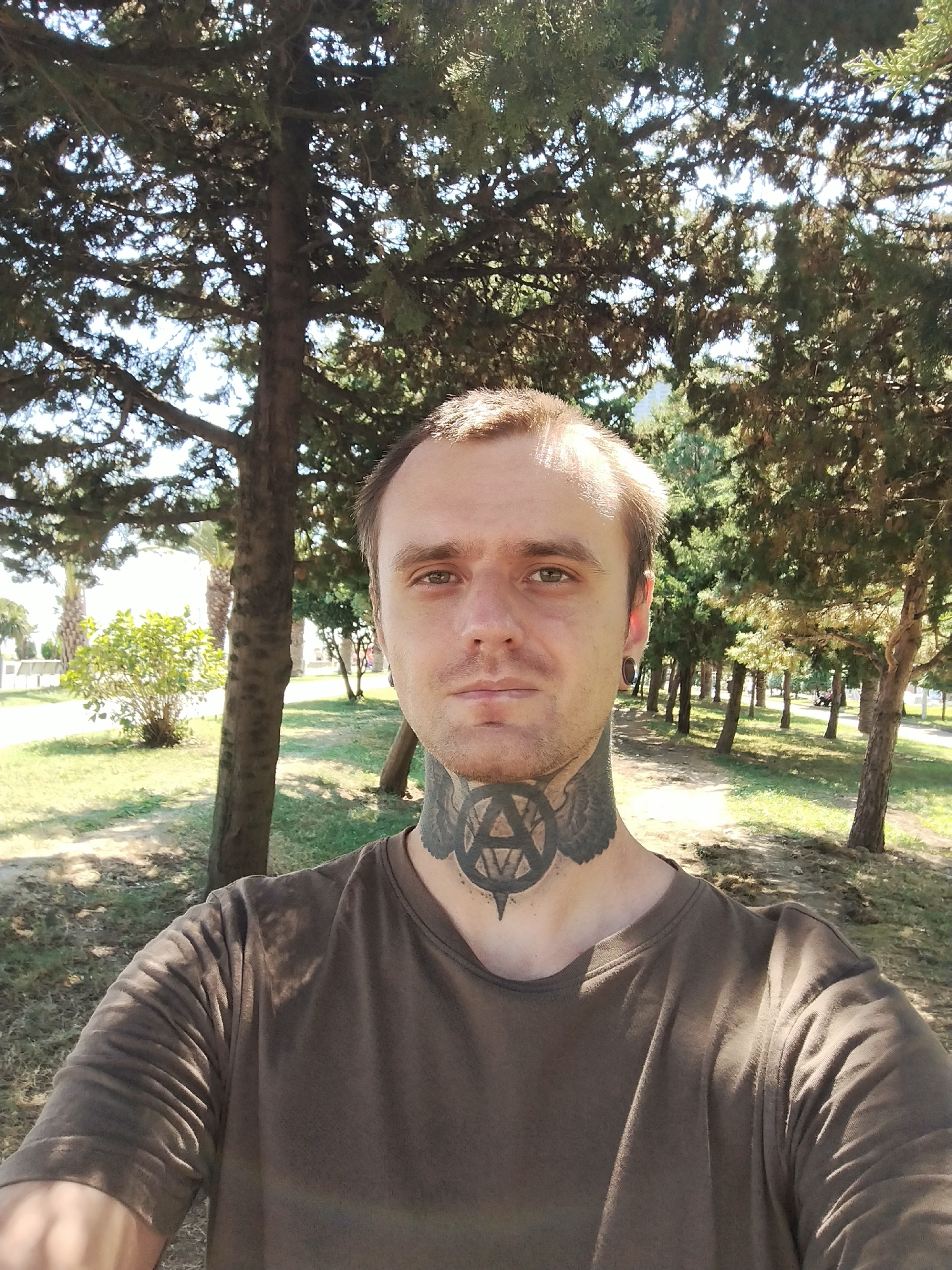 The image is a selfie taken outdoors in Batumi, Georgia. The subject is a white male with short brown hair, light skin, and a tattoo around his neck. He is wearing a brown t-shirt. The background features several trees with green foliage and a grassy area. The lighting suggests it is daytime, with sunlight filtering through the trees. The environment appears to be a park or a similar public outdoor space.
