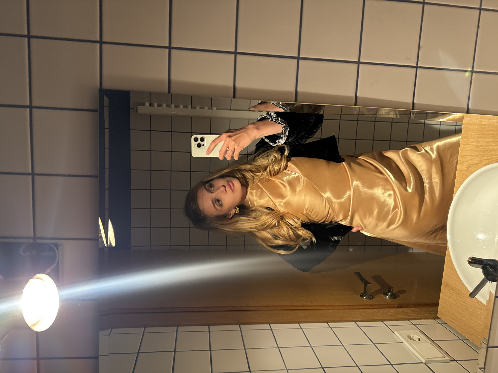 The image shows a selfie taken in a bathroom, likely in Tbilisi, Georgia. A young woman with blonde hair and fair skin is reflected in a mirror. She's holding an iPhone in her right hand. She's wearing a gold satin dress and a black velvet jacket with decorative cuffs. The background features a tiled wall, a sink, and a light fixture. The tiles are square and white with dark grout. The sink is white and appears to be part of a wooden vanity. There is an electrical outlet on the wall near the floor.