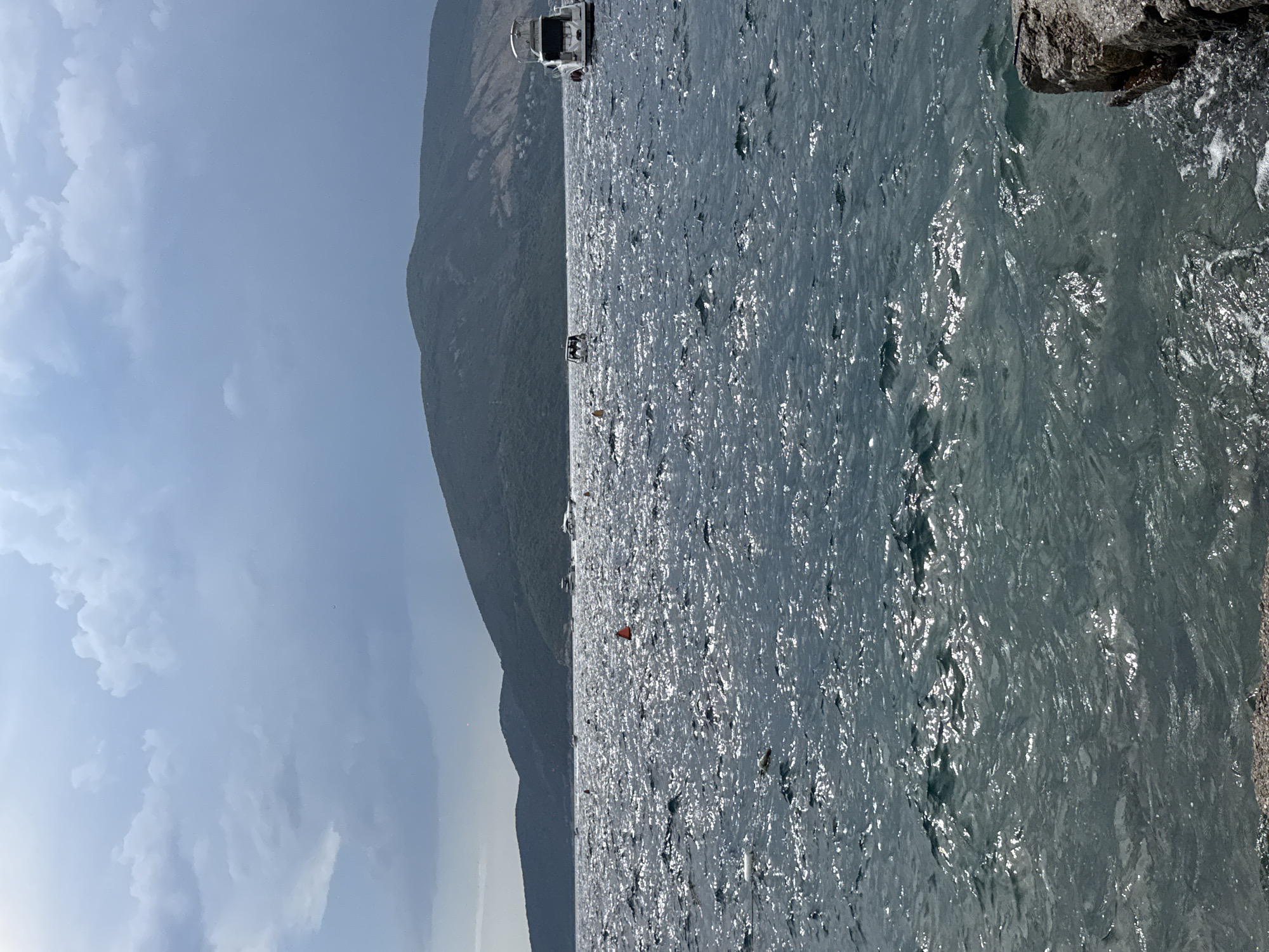 The image depicts a body of water with choppy, sunlit waves. In the distance, a range of verdant, mountainous terrain forms the horizon. Several boats are visible on the water. On the right side of the frame, a white motor yacht is docked or anchored. Towards the center, a smaller white boat is also present. Further to the left, there are a few buoys and other small vessels. The sky above is partly cloudy with some darker, storm-like clouds on the left, contrasting with brighter areas on the right. The foreground shows a rocky shoreline or pier. The overall scene suggests a coastal or lakeside environment under variable weather conditions.