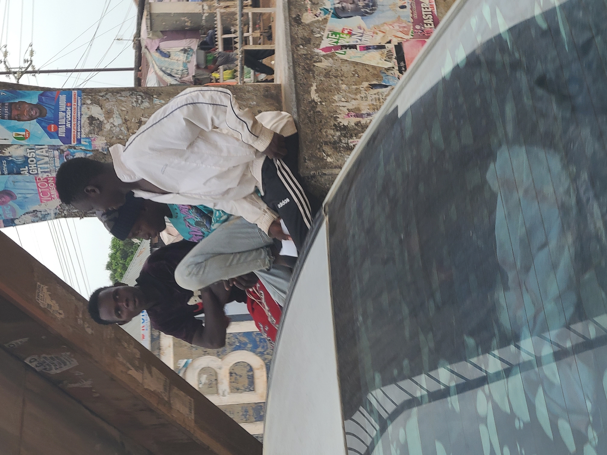 The image captures an outdoor street scene in Gura Top, Nigeria, featuring several individuals and elements of an urban environment. The perspective is slightly rotated.
In the foreground, a dark-colored vehicle is partially visible on the right, showing a reflective side window. A large wooden beam or structure runs diagonally across the lower left.
The main subjects are three men gathered near a wall covered with numerous posters and flyers. One man, in a white long-sleeved shirt and white track pants with black stripes and an "adidas" logo, is bending over, seemingly looking down or interacting with something off-frame. Next to him, another person in a patterned top is partially visible. Further to the left, a man in a dark red short-sleeved shirt and light-colored jeans looks directly towards the camera.
The wall behind the individuals is extensively plastered with various notices. Prominently visible are political campaign posters, including one for "AYUBA DALYOP MANDUNG (AYUDEH)" running for "MEMBER PLATEAU STATE HOUSE OF ASSEMBLY JOS SOUTH CONSTITUENCY," bearing the "APC" party logo. Other posters mention "GHOST FESTIVAL," "SAMSON GREATER," and "EASTER."
The background shows parts of buildings and what appears to be vegetation under a clear, bright sky, indicating daytime. The overall atmosphere suggests a typical day in a bustling roadside area. No strong emotions are evident from the subjects; their expressions are mostly neutral.