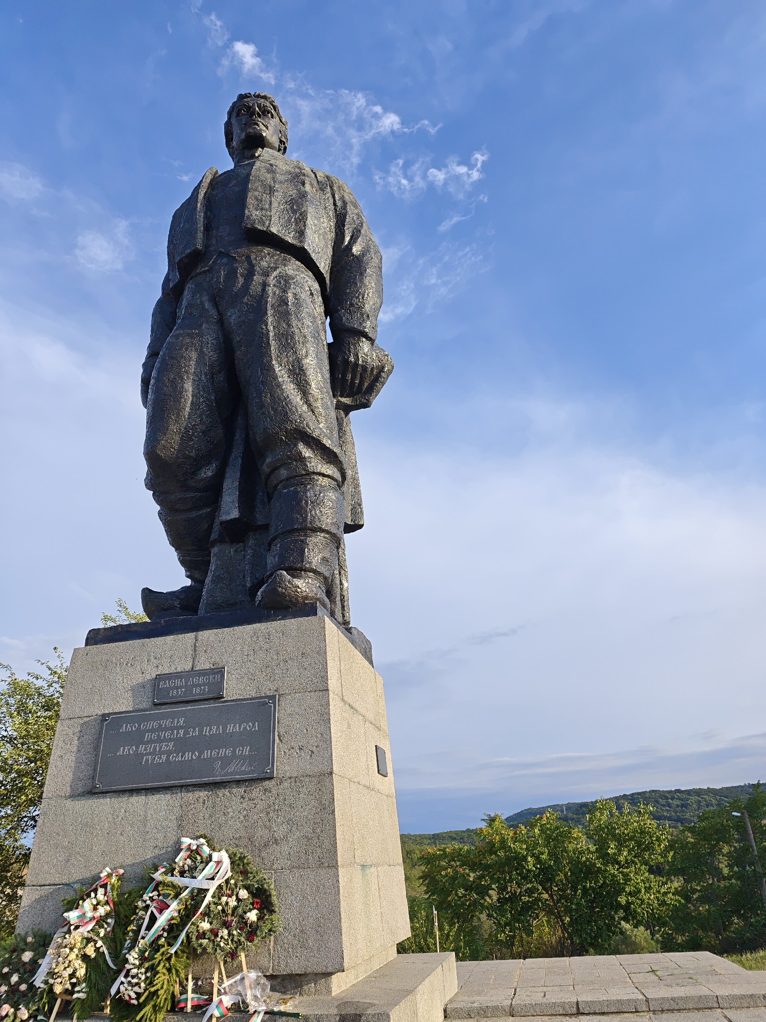 This image shows a statue of Vasil Levski, a national hero of Bulgaria, located in the city of Lovech. The statue is made of dark metal and depicts Levski standing tall, looking upwards. He is dressed in traditional Bulgarian attire, including a jacket, trousers, and boots. The statue is placed on a large, granite pedestal.
On the pedestal, there are two plaques. The upper plaque has the name "ВАСИЛ ЛЕВСКИ" (Vasil Levski) and the years "1837 - 1873". The lower plaque contains a quote in Bulgarian, which translates to: "...If I die, I will die for Bulgaria, and then a nation will rejoice." This quote is attributed to V. Levski.
In front of the pedestal, there are several wreaths of flowers, adorned with ribbons in the Bulgarian national colors (white, green, and red). These appear to be tributes placed at the monument.
The background of the image shows a bright blue sky with scattered white clouds. To the right of the statue, there are trees and a hillside covered in green vegetation, suggesting an outdoor, elevated location. The overall lighting indicates it is daytime, possibly late afternoon, given the angle of the sunlight.