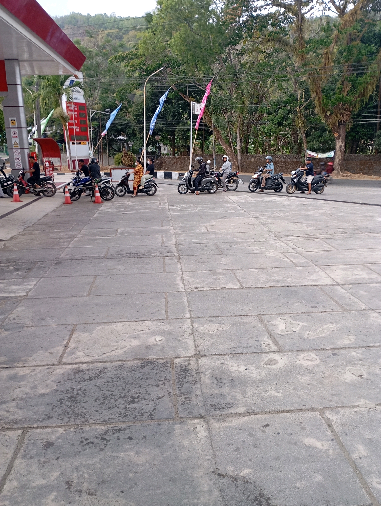 The image captures a street scene at a gas station in Menadi, Indonesia. A red and white canopy of a gas station is visible on the left. Multiple motorbikes are parked in a line, with several individuals on or near them. On the left, a person in a red shirt is on a motorbike. Next to them, two men in dark clothing are on a motorbike, one wearing a helmet. Further down, a woman in a patterned dress is seated on a motorbike, and another person in dark clothing is also on a motorbike. The next two motorbikes have riders wearing helmets, one in a blue shirt and the other in a dark shirt and shorts. A red cone is placed near the front of some of the motorbikes. Tall trees and a stone wall form the background, with power lines visible above. Two blue flags and one pink flag are attached to poles on the right side of the frame. The ground is paved with large, grey rectangular stones.