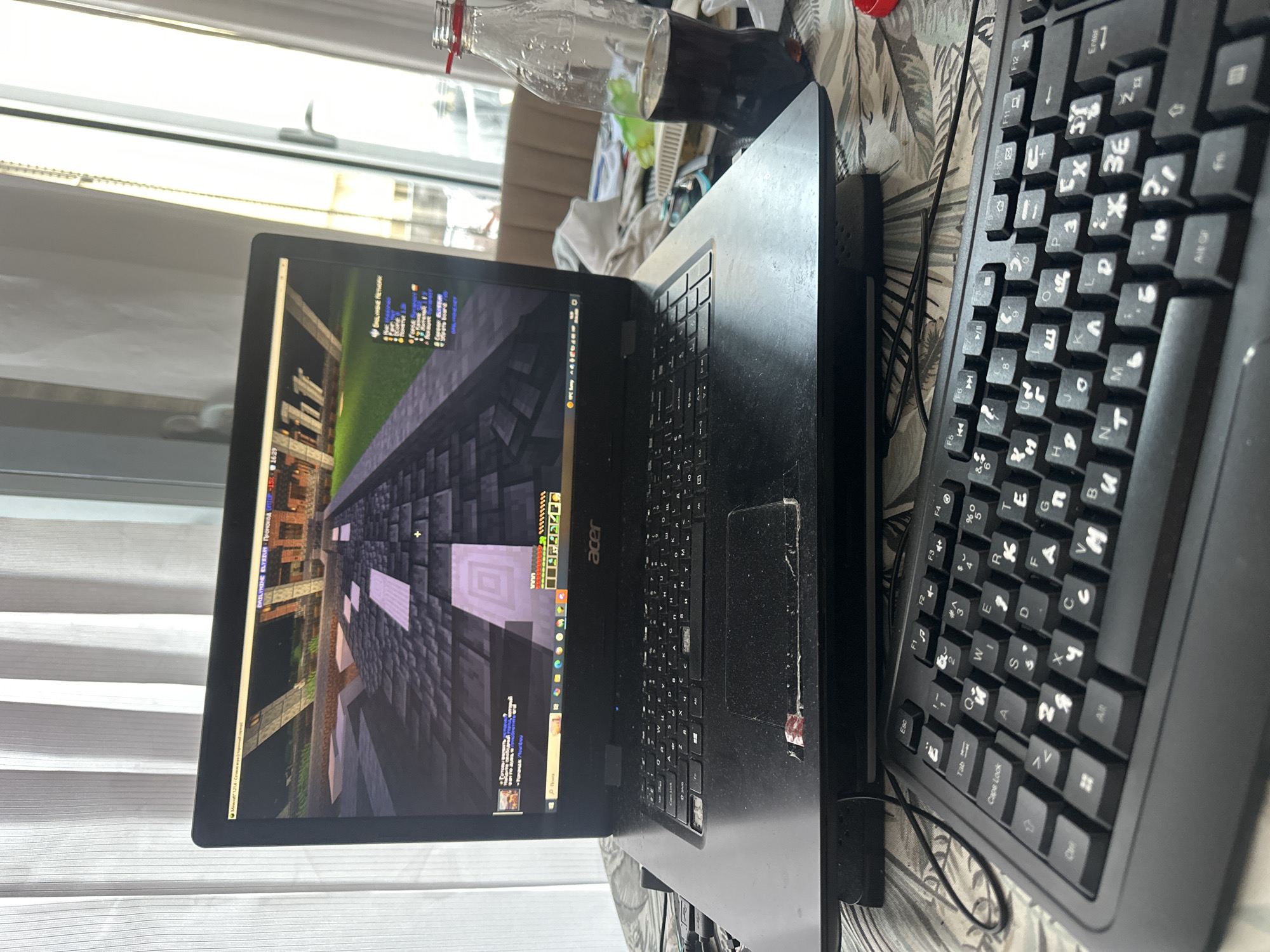 The image captures a casual indoor scene in Budva, Montenegro, likely during the daytime, focusing on a laptop setup. The primary subjects are an open black Acer laptop and an external black keyboard, positioned on a table covered with a patterned cloth.
The laptop screen displays a video game, resembling Minecraft, characterized by a pixelated, block-based world. On the game's user interface, text such as "REAL MINE Network" and a list of game modes or servers including "Forget", "Bounty", "Servers", "Creative", "SkyWars", "Bedwars", and "Online React" are visible. In the bottom left, "Minecraft 1.14" is displayed, alongside a promotion for "THE ELYSIUM Промокод" (promo code). The top right corner of the screen shows game statistics like "FPS", "Ping", and "Time".
An external black keyboard with both Latin and Cyrillic characters on the keys is placed next to the laptop, suggesting its use in a region where both alphabets are common, consistent with the Montenegro location context. Keys like "Tab", "Caps Lock", "Enter", and various function keys (F1-F12) are discernible, along with letters such as "Q", "W", "E", "R", "A", "S", "D", "F", and Cyrillic counterparts like "Ц", "У", "К", "Е", "Н", "Г", "Ф", "Ы", "В", "А", "П", "Р", "О", "Л", "Д", "Ж", "Э", "Я", "Ч", "С", "М", "И", "Т", "Ь", "Б", "Ю".
To the right of the laptop, a partially full, clear plastic bottle containing a dark liquid, identifiable as Coca-Cola from a visible label fragment, stands. The background features a window with white blinds, allowing natural light into the room. The laptop and keyboard show some signs of dust on their surfaces. No people are visible, and emotions cannot be discerned.
