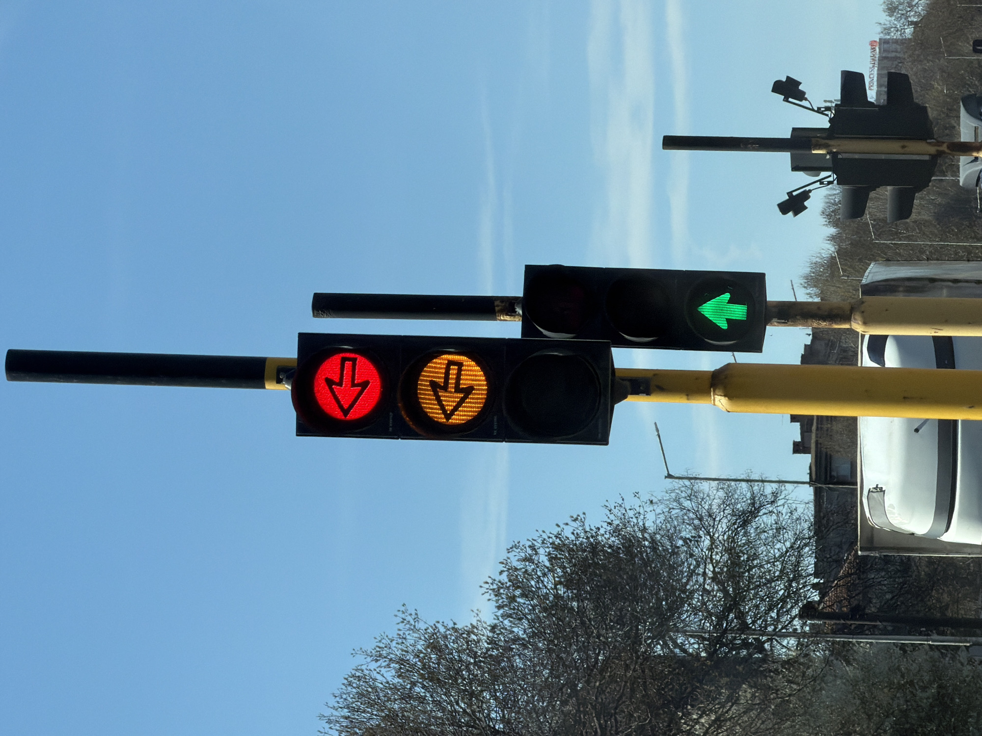 The image shows two traffic lights against a bright blue sky with wispy clouds. The lower traffic light displays a red arrow pointing down and an orange arrow pointing down, while the upper traffic light displays a green arrow pointing to the right. The traffic lights are attached to yellow poles. Bare tree branches are visible in the lower right of the frame. A vehicle and buildings are partially visible in the right of the frame. Based on the style of traffic lights and urban environment, this is likely located in Sofia, Bulgaria.