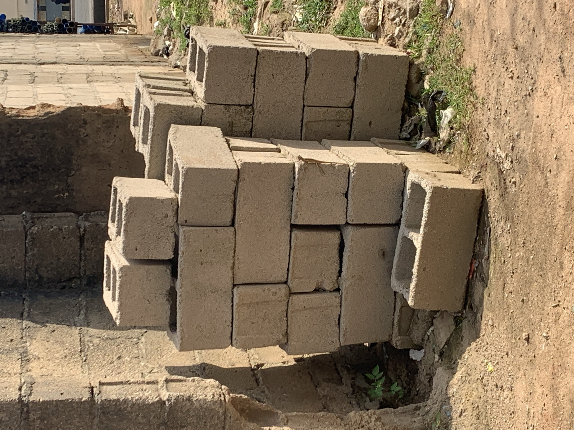 This image shows a pile of concrete blocks stacked on the ground. The blocks are of various sizes and appear to be standard construction blocks. Some of the blocks have holes running through them. The pile is situated on a dirt and gravel surface, with some patches of grass and weeds growing around it. In the background, there is a hint of a building with some pipes or containers stacked in front of it. The lighting suggests it is daytime, with shadows cast on the ground, indicating a sunny environment. The overall scene appears to be an outdoor area, possibly a construction site or storage area for building materials. There is no visible text in the image. No people are present. The scene is static, with no activity taking place.