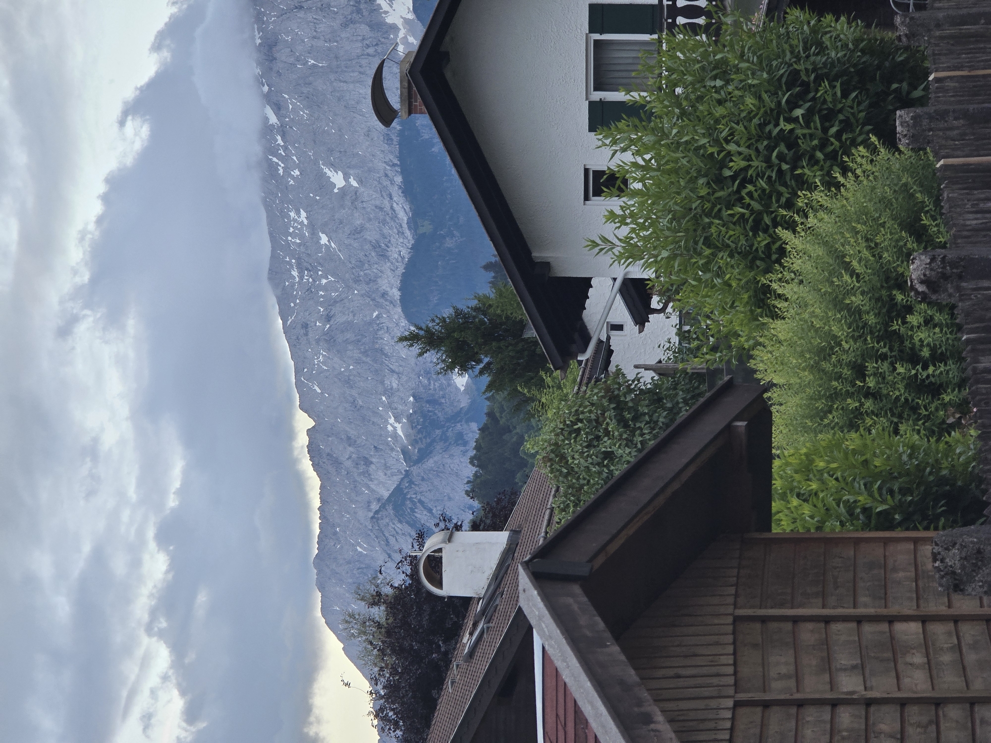 This image depicts a residential and natural landscape in Garmisch-Partenkirchen, Germany. The foreground features a weathered wooden fence with grey, coarse posts. Immediately behind the fence, dense, vibrant green bushes and shrubs grow, partially obscuring the lower sections of several structures. On the far left, a close-up shows the sloped, dark brown roof and wooden plank siding of a shed-like building.
In the midground, traditional German houses are visible. One prominent house on the right features white stucco walls, a dark sloped roof, and dark green closed shutters on its windows. A chimney with a dark, curved cap is situated on this roof. To the left, another house with a tiled brown roof and a distinctive white, rounded chimney with an open arch is partially visible. Various trees and substantial green foliage, including a dark reddish-purple tree and a tall conifer, are interspersed among the houses, providing natural cover.
The background is dominated by a majestic, rugged mountain range, consistent with the Bavarian Alps. The grey rock faces are dotted with numerous patches of white snow, indicating a significant elevation or recent snowfall. The lower slopes of the mountains are covered with dark green forests. Overcast conditions prevail, with a thick layer of grey and white clouds partially obscuring the mountain peaks and filling the sky above, creating a diffused light across the scene. No individuals or specific actions are depicted.