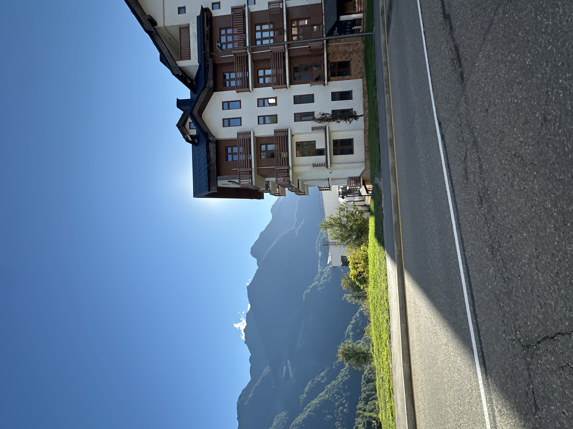 The image displays a contemporary apartment building with a light-colored facade, accented by dark brown wooden elements and balconies. The building appears to be situated in a mountainous region, as indicated by the expansive, tree-covered mountains that dominate the background under a clear blue sky. The sun is shining, suggesting it is daytime. A paved road runs alongside the building and the green, grassy verge. The scene suggests a tranquil, possibly residential or resort-like environment in a natural setting. No people are visible in the image, and there is no discernible text. The orientation of the image is rotated 90 degrees counter-clockwise from the standard upright view.