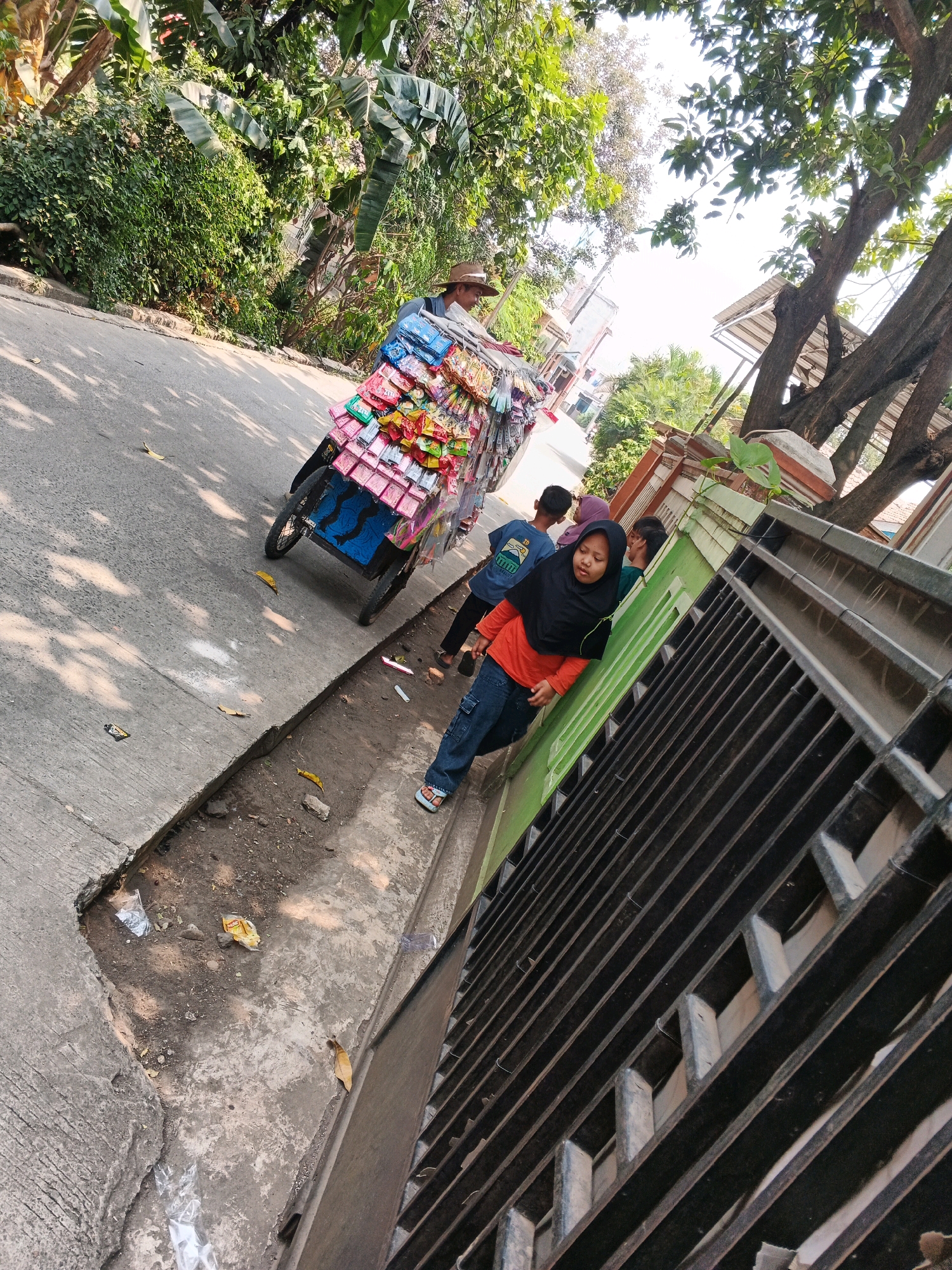 In the city of Kadu, Indonesia, a street scene unfolds with a vendor pushing a cart laden with colorful snacks along a paved road. The cart, a three-wheeled vehicle, is filled with rows of individually packaged snacks. To the right of the road, a low green wall runs parallel to the street, behind which is a black metal fence. Several individuals are present. A person wearing a hat, presumably the vendor, is partially visible behind the cart. Closer to the fence, a young person in a black hijab and an orange shirt is walking, looking down. To their left, two other young people, one in a blue shirt and the other wearing a purple hijab, are also walking away from the cart. The environment is characterized by lush green foliage and trees lining the street, with sunlight casting dappled shadows on the asphalt. The ground near the wall and fence is littered with some debris, including plastic wrappers and fallen leaves, suggesting a typical urban streetscape.