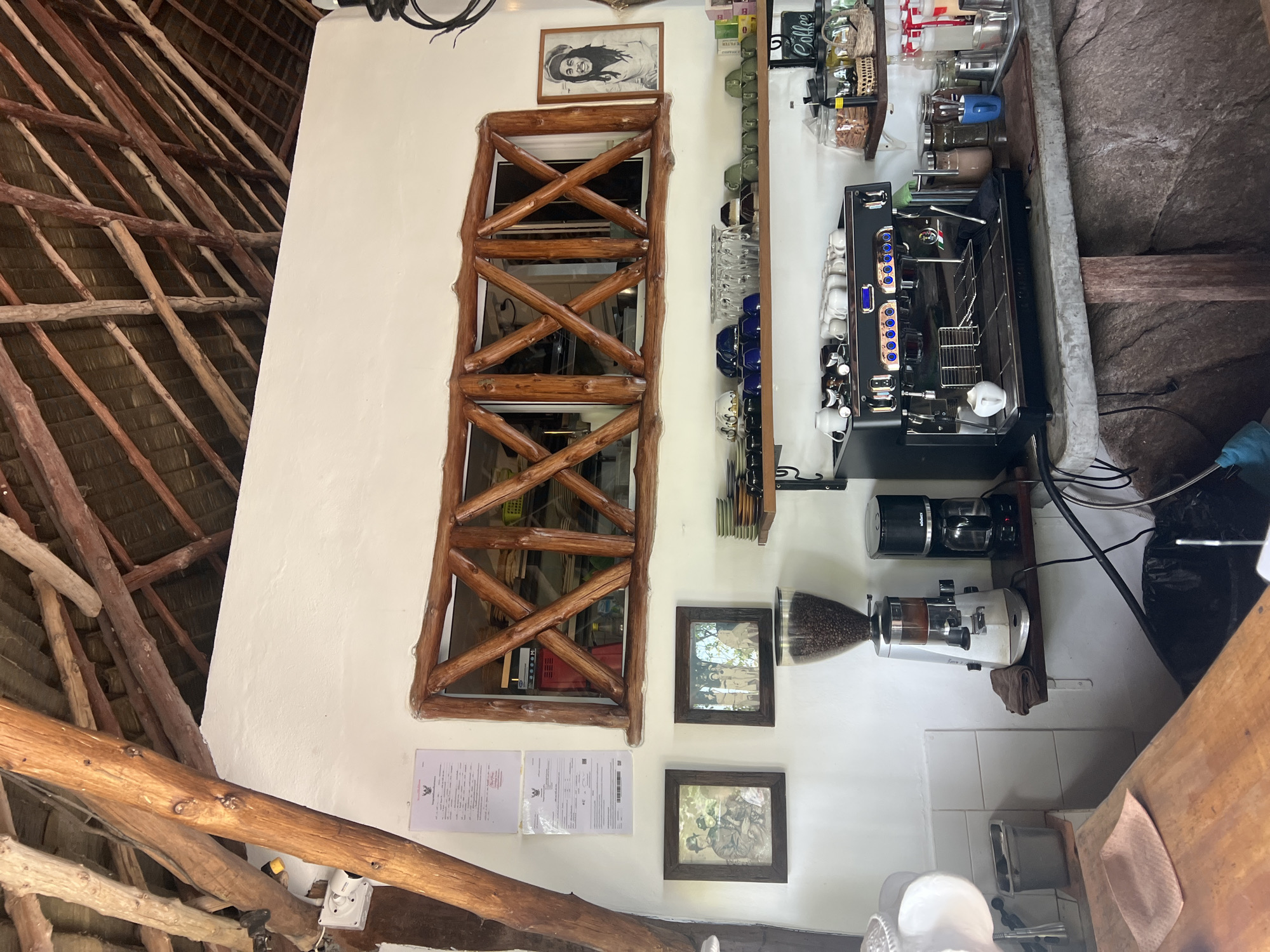 This image depicts the interior of a cafe with a rustic, natural ambiance, likely located in Baan Chaweng Noi, Thailand. The ceiling is constructed with natural wooden beams and thatch. The walls are white, with a large, artfully crafted wooden frame above a window that looks into what appears to be the cafe's preparation area. On the wall, two framed documents are visible, along with two framed vintage photographs.
A coffee grinder with a hopper full of coffee beans stands on a shelf next to a drip coffee maker. Below these, a professional-grade espresso machine is prominently displayed on a concrete counter. Shelves above and to the right are stocked with various cups, saucers, glasses, and jars of coffee-related items, including a jar labeled "Coffee". To the right of the window, a framed portrait of Bob Marley is hanging on the wall.
The scene appears to be a service area of a cafe, possibly in the late morning or early afternoon, given the lighting. No people are directly visible in the main focus of the image, but the presence of a coffee machine and associated equipment suggests a functioning establishment. The overall environment suggests a relaxed, tropical setting.