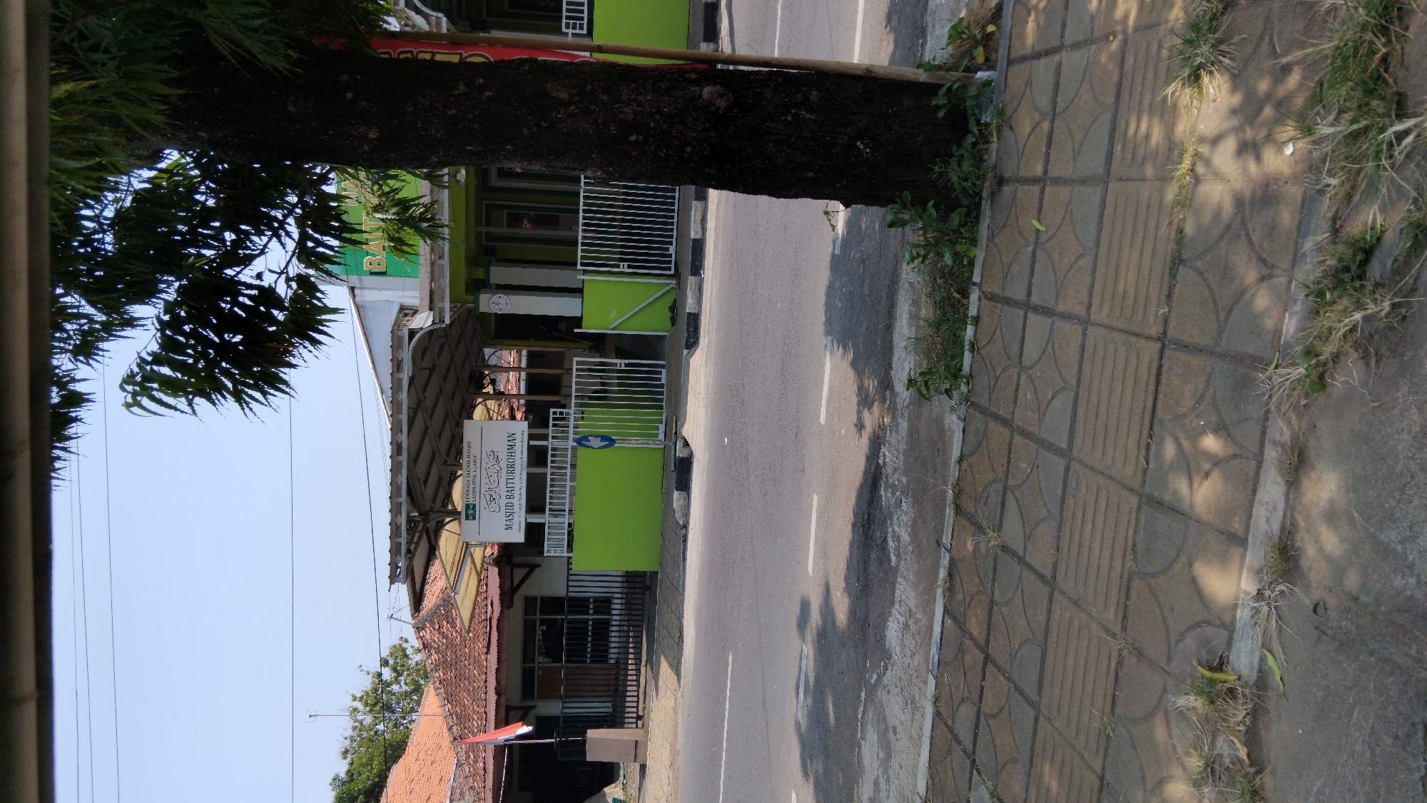 The image shows a street-level view of Masjid Baiturrohman in Proyonangg, Indonesia, under a clear blue sky. The mosque's facade is painted green, with a white fence enclosing its entrance. A sign above the entrance reads "LEMBAGA YAYASAN MASJID KABUPATEN ILL... ILAM." and "MASJID BAITURROHMAN". A tall, dark tree with dense foliage stands to the right of the mosque, partially obscuring the view. The street in front of the mosque is paved and has white lane markings. A sidewalk with patterned tiles runs along the side of the street, with grass growing between the tiles and along the edge. A red and white Indonesian flag is visible on a flagpole to the left of the mosque. The scene appears to be quiet and serene, with no people visible.