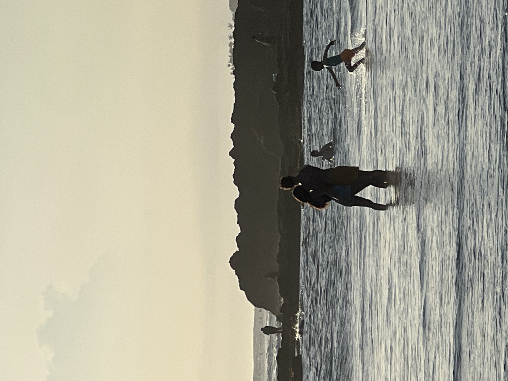 The image depicts a seaside scene with several individuals in the water against a backdrop of a hilly landmass. One person is running towards the open sea, while a pair appears to be playfully wrestling. In the distance, a figure stands at the shoreline. The location is likely North Kuta, Indonesia. The water exhibits a wavy texture, reflecting the ambient light.