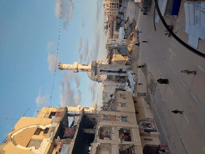 This image depicts a scene of extensive urban destruction in Khan Yunis, Palestinian Territory. The focal point is the heavily damaged Great Mosque of Khan Yunis (Al-Hamza Mosque) in the mid-background, identifiable by its partially intact minaret and dome, surrounded by significant structural collapse and debris. To the left, a multi-story building, likely residential or commercial, exhibits severe damage with large sections of its facade missing, revealing interior rooms and exposed floor levels. Laundry hangs from damaged balconies on this structure. Further buildings in the background also show varying degrees of damage, with some reduced to rubble piles.
The asphalt street, with visible white road markings, traverses the scene. It is largely clear of major debris, but significant piles of rubble and fractured building materials are visible along its edges and around the damaged structures. The sky overhead is light blue with scattered white clouds.
Approximately 15-20 individuals are present in the scene. In the central foreground, two figures in dark, flowing garments walk on the road, with another lone individual in lighter attire positioned further ahead. Several other people, including some children, are scattered across the street and sidewalks, generally appearing to be walking through the area. A dark cable or wire extends across the upper right foreground, partially obscuring a view of a makeshift structure covered in white sheeting on the far right.