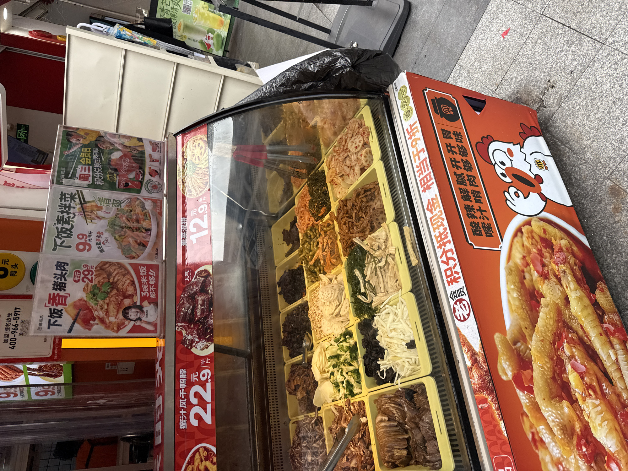 A street vendor displays a variety of prepared food items in a refrigerated glass case. The case is filled with yellow rectangular containers holding different kinds of ingredients, including what appear to be thinly sliced meats, vegetables, and possibly some preserved items. Tongs are visible resting on some of the food. Above the display case, various signs advertise the food. One sign features a picture of a young girl and braised pork, with Chinese text. Another sign has a graphic of people celebrating and mentions Coca-Cola and the World Cup. A prominent sign at the bottom of the display advertises a dish with chicken feet, with the text "酸辣解腻开胃 唆汁肉够味够鲜" (Sour and spicy, refreshing and appetizing, soupy and flavorful chicken is delicious and fresh). The overall scene suggests a food stall or small restaurant in an urban environment. The image appears to be taken in Haizhu District, China, based on the information provided.