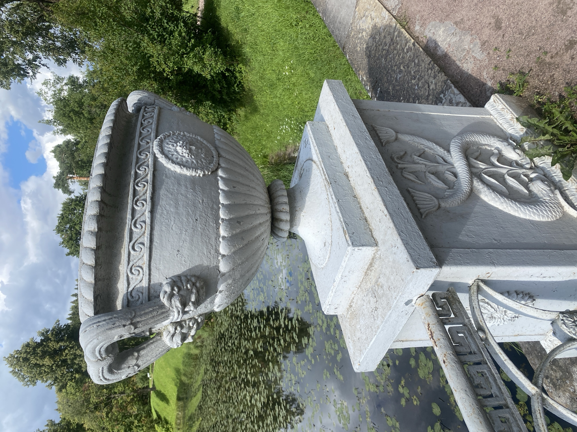 An ornate, weathered, white stone urn sits atop a square pedestal, which is part of a stone balustrade. The urn has detailed carvings, including lion heads supporting its handles and a circular relief of a face on its side. The pedestal also features relief carvings of floral motifs and a serpent. The balustrade extends to the left, with a metal railing in front of it, displaying a Greek key pattern.
Behind the urn, a dark body of water is visible, covered with numerous lily pads. The water reflects the sky and the surrounding greenery. The background consists of lush green grass, dense trees, and a cloudy blue sky. The scene appears to be in a park setting. This location is the Pavlovsk Park, a UNESCO World Heritage Site in Pavlovsk, Russia, specifically on the picturesque bridge over the Slavyanka River. There are no people present in the image.
