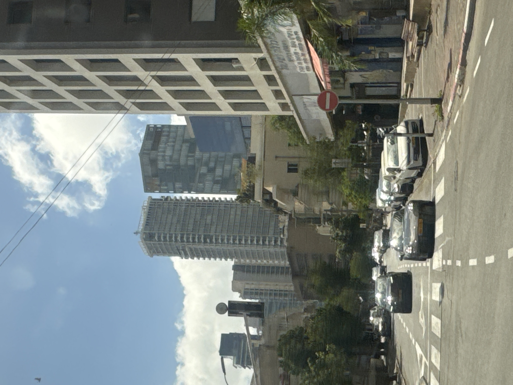 The image shows a street scene in Tel Aviv, Israel. On the right side of the frame is a street with parked cars. The street has white dashed lines indicating traffic lanes. The background features several tall buildings, including skyscrapers. There are trees and greenery interspersed among the buildings and along the street. The sky is visible in the background, with some clouds. There is a "Do Not Enter" sign visible.