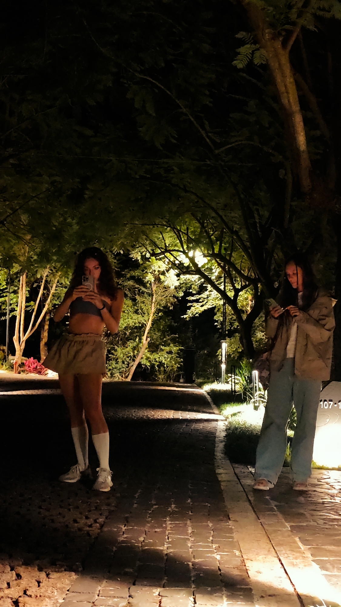 This nighttime image, set in Mu Cang Chai, Vietnam, shows two young women on a well-lit, brick-paved path bordered by dense, illuminated trees and foliage. The woman on the left, with long, dark curly hair, wears a light crop top, a light ruffled skirt, white knee-high socks, and athletic shoes, holding a smartphone in front of her face, apparently taking a photograph or video. The woman on the right, with long dark hair, wears a light jacket, light blue wide-leg jeans, and light slip-on shoes, looking down at a smartphone in her hands. Several vertical path lights illuminate the greenery from below, casting a warm glow. A partially visible sign to the right displays "107-1".