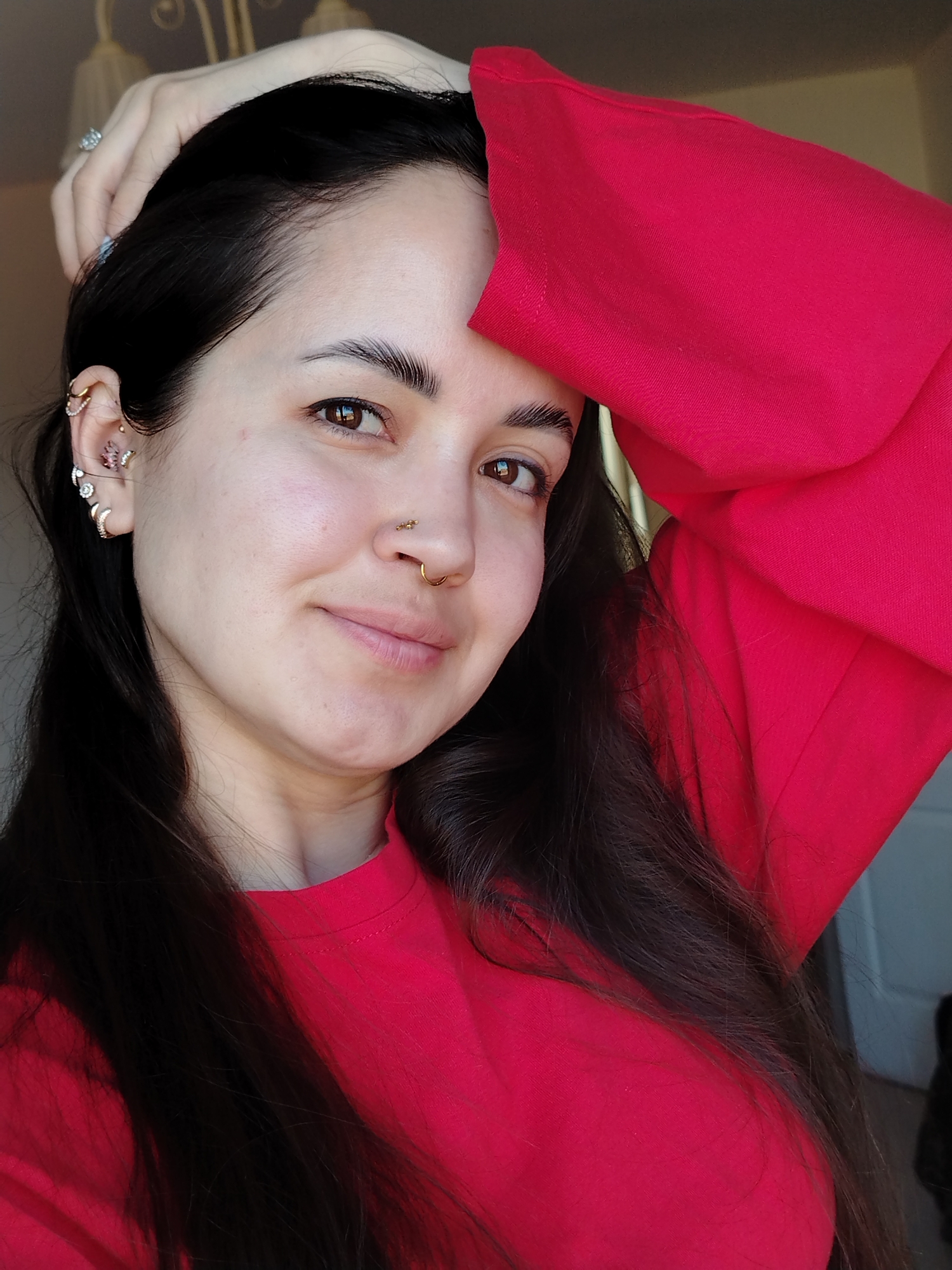 A close-up, brightly lit portrait captures a young woman looking directly at the camera with a gentle smile. She has long, dark brown hair, which she holds back with her left hand placed on top of her head, revealing a ring on her ring finger. Her dark eyebrows are well-defined, and her brown eyes reflect the light, suggesting a daytime setting.
Her face is clear, and she has several facial piercings: multiple piercings adorn her left ear, including various studs and hoops, one of which features a pink, floral or butterfly-like design. She also has two nose piercings – a small gold stud on the side of her nose and a more prominent gold hoop septum ring.
She is wearing a vibrant red, long-sleeved top or sweatshirt, which occupies a significant portion of the lower and right side of the frame. The fabric appears soft with a visible texture. The background is softly blurred, showing hints of an indoor setting, possibly a room, with a light-colored wall or door on the right and an out-of-focus light fixture above her head. The overall mood is pleasant and casual. While the image itself doesn't offer specific visual cues, the provided context indicates the location is Noviy, Russia. There is no visible text in the image.