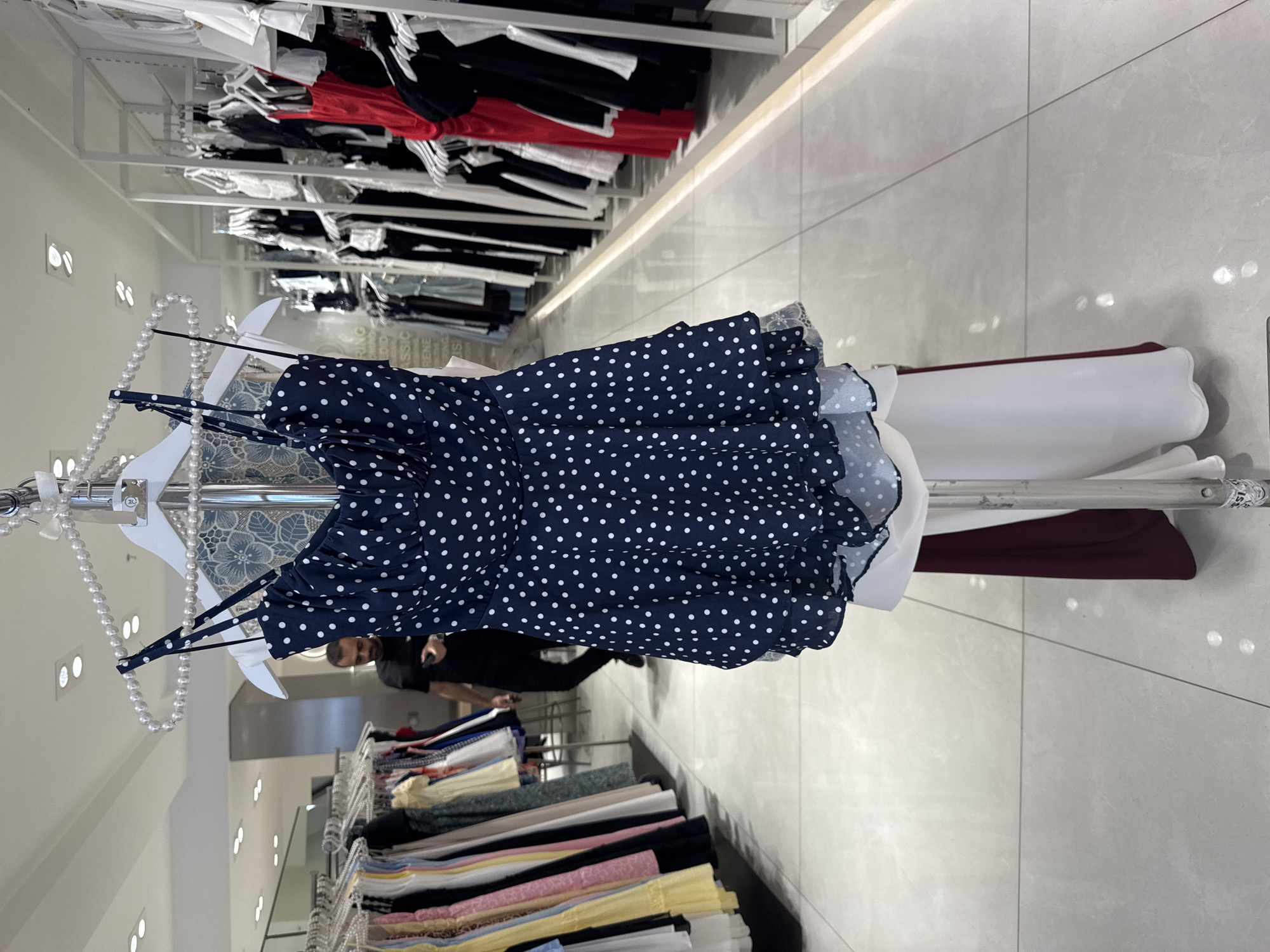 The image shows a dark blue polka dot dress hanging on a hanger in a clothing store. The dress has thin straps and a ruffled hem. In the background, a man is walking through the store, and racks of clothing are visible on either side. The store appears to be a modern boutique with white walls and tiled floors. The overall scene suggests a commercial environment focused on fashion retail.