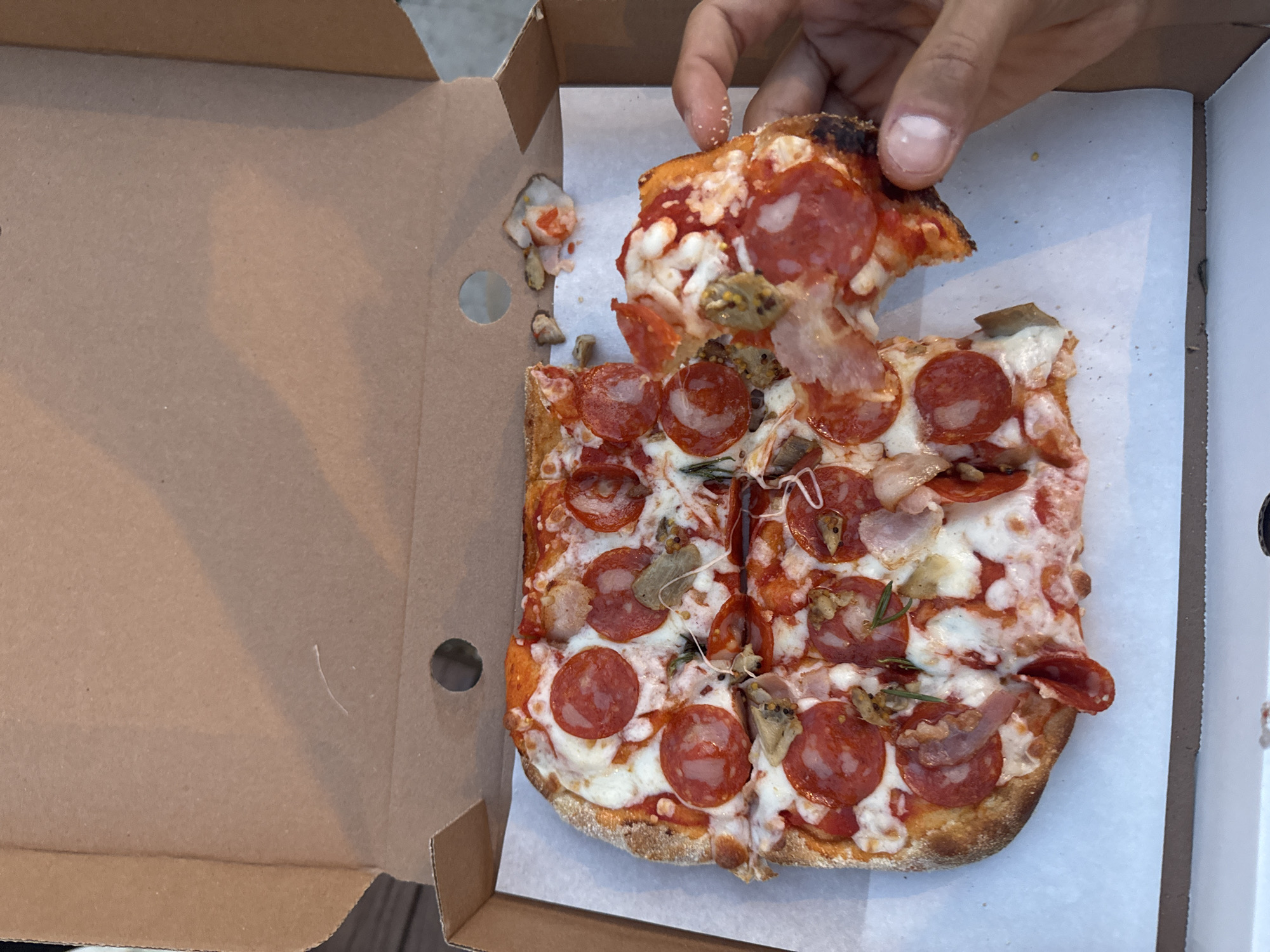 A rectangular pizza in a cardboard box is shown from an overhead perspective. A dark-skinned hand is taking a slice from the pizza, partially lifting it from the box. The pizza is topped with pepperoni, mozzarella, what appears to be cooked mushrooms, and herbs, likely rosemary. The box is open, revealing the pizza sitting on white paper. The edges of the box are visible, with a circular cutout on the left side. The environment suggests an informal setting, possibly outdoors given the natural lighting. There is no information in the image to indicate the location of Moscow, Russia.