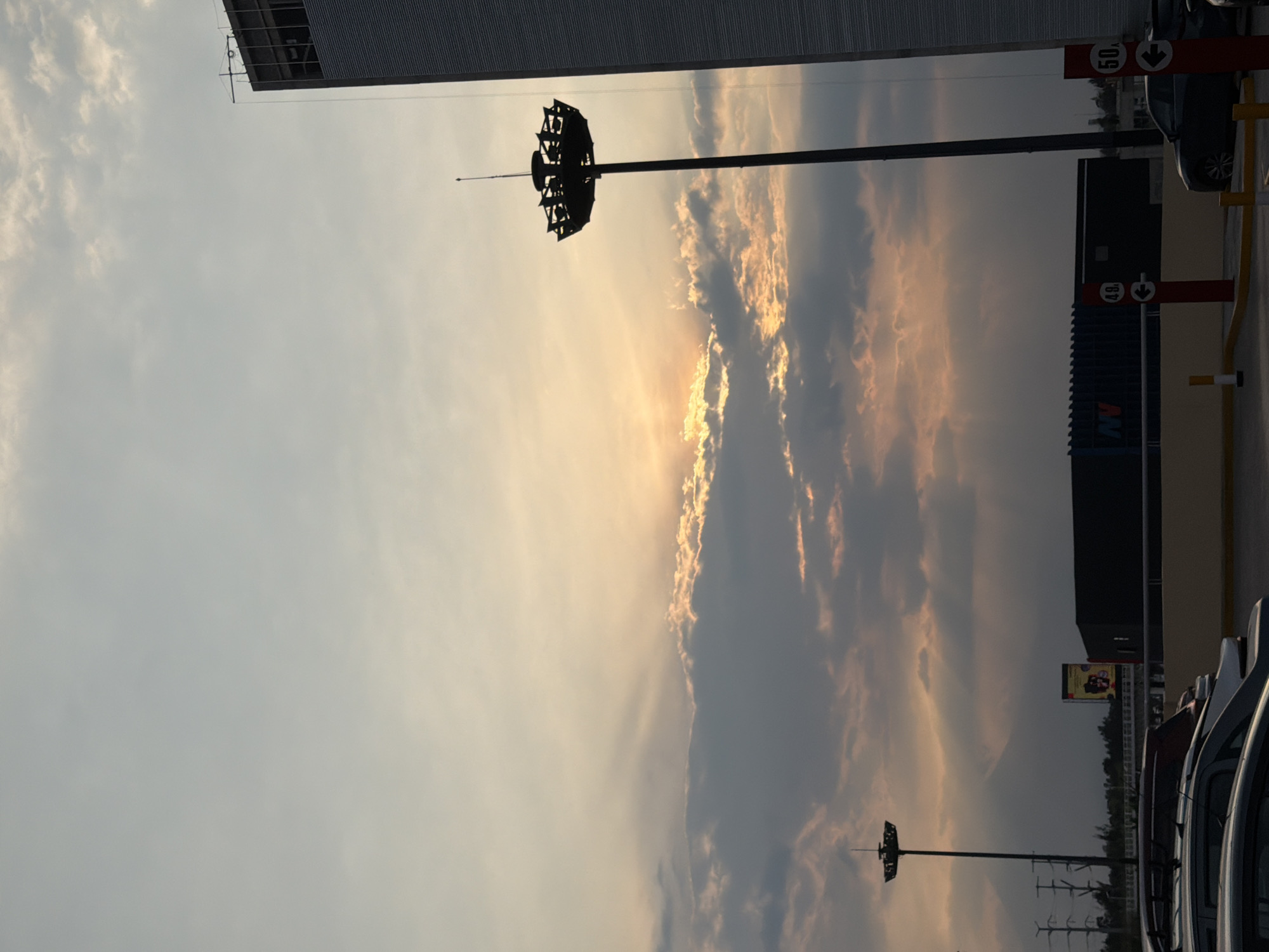 The image, taken in an urban setting, possibly a parking lot or commercial area, shows a sky at dusk with dramatic clouds partially obscuring the setting sun. The sky is light in color transitioning to darker shades due to the cloud cover. Tall stadium-style light poles with complex lamp arrays at the top are silhouetted against the sky. A large building with a grey facade is visible at the top of the frame, partially obstructing the sky. Cars are parked on the right side of the frame. A poster or advertisement is also visible in the background. Road signs are installed near cars.