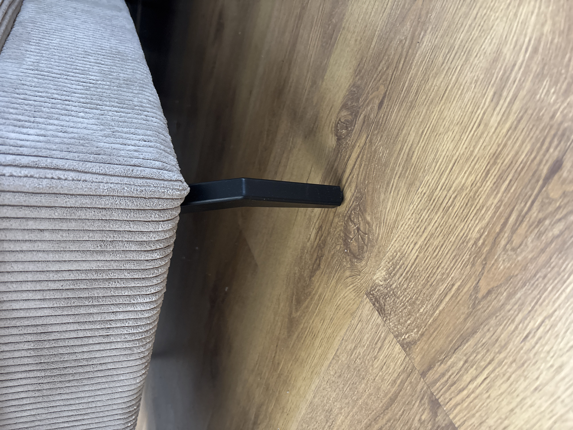 The image is a close-up, low-angle shot of a single, black, angled furniture leg supporting a beige, corduroy-textured sofa. The leg is made of metal and is positioned on a light brown laminate wood floor. The floor exhibits a wood grain pattern. The background is dark and out of focus, hinting at the interior of a room. No people or other objects are visible. The location is Unknown, Unknown.