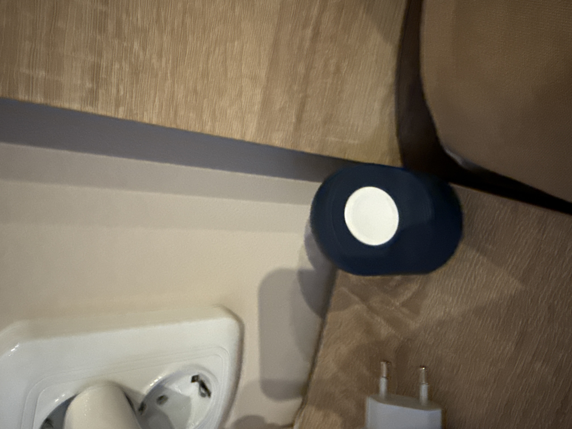The image shows an electrical outlet with a European plug inserted, positioned near a piece of furniture. A blue and white circular object is placed on the furniture adjacent to the outlet. The furniture has a light brown texture. The location is indiscernible from the image. The city of Lovech, Bulgaria cannot be confirmed by visual evidence alone.