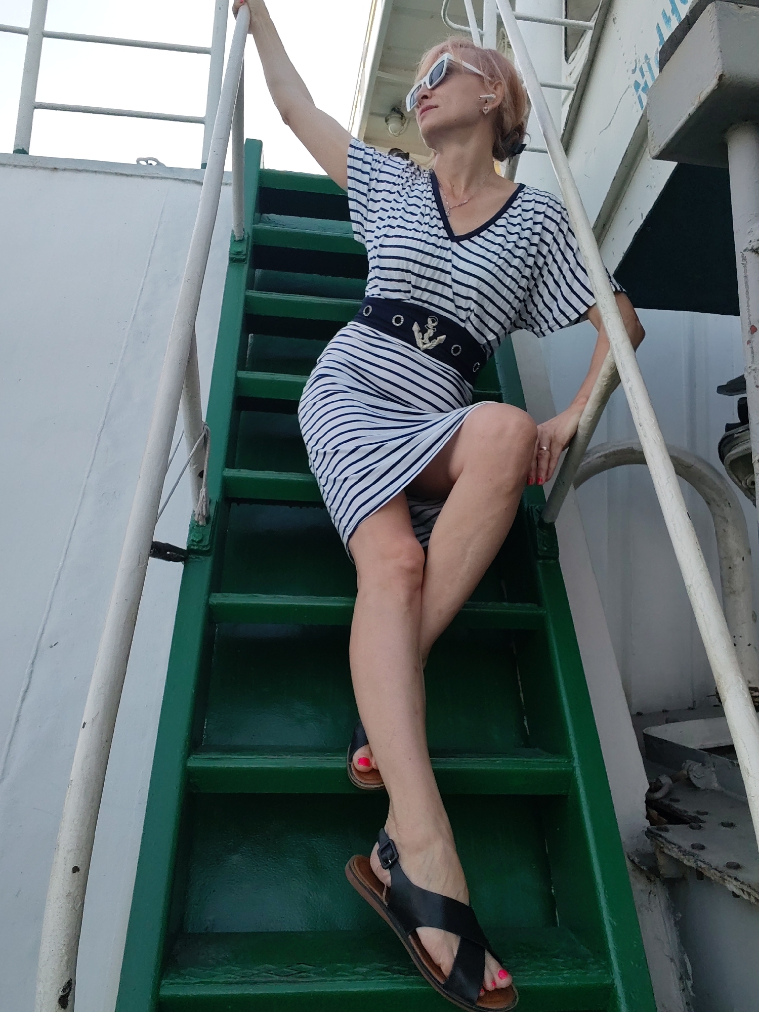 An adult female with short, pink hair and white sunglasses is depicted on the green metal stairs of a vessel. She is wearing a white and black striped dress with a black belt featuring an anchor emblem. Her left arm is raised, grasping a white railing, while her right hand rests on the railing. She is positioned on the stairs, with her legs bent. The surrounding environment appears to be the exterior of a ship, with white walls and metal structures visible. The image is taken from a low angle, looking up at the woman. The location is identified as Shchetinino, Russia.