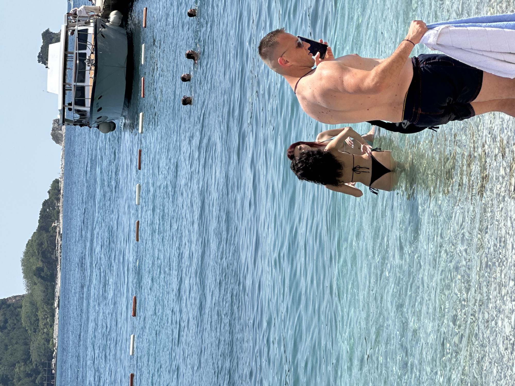 This is a candid, eye-level, outdoor, medium shot captured on a sunny day at a beach in Budva, Montenegro. In the foreground, a muscular man wearing black swim trunks and sunglasses is holding a cell phone and a light blue towel. He appears to be looking at his phone. Behind him, two women are standing in clear, shallow water. One woman with dark hair wears a black bikini, and the other, with red hair, wears a light-colored bikini. In the background, several people are swimming in the ocean, and a white boat is docked next to a pier. The water is calm and a light blue color, with some ripples on the surface. The shoreline is visible with a pebble beach. In the far distance, a green, treed hill rises from the water, and a few small rocky islands are scattered in the sea. A line of orange and white buoys demarcates an area of the water.