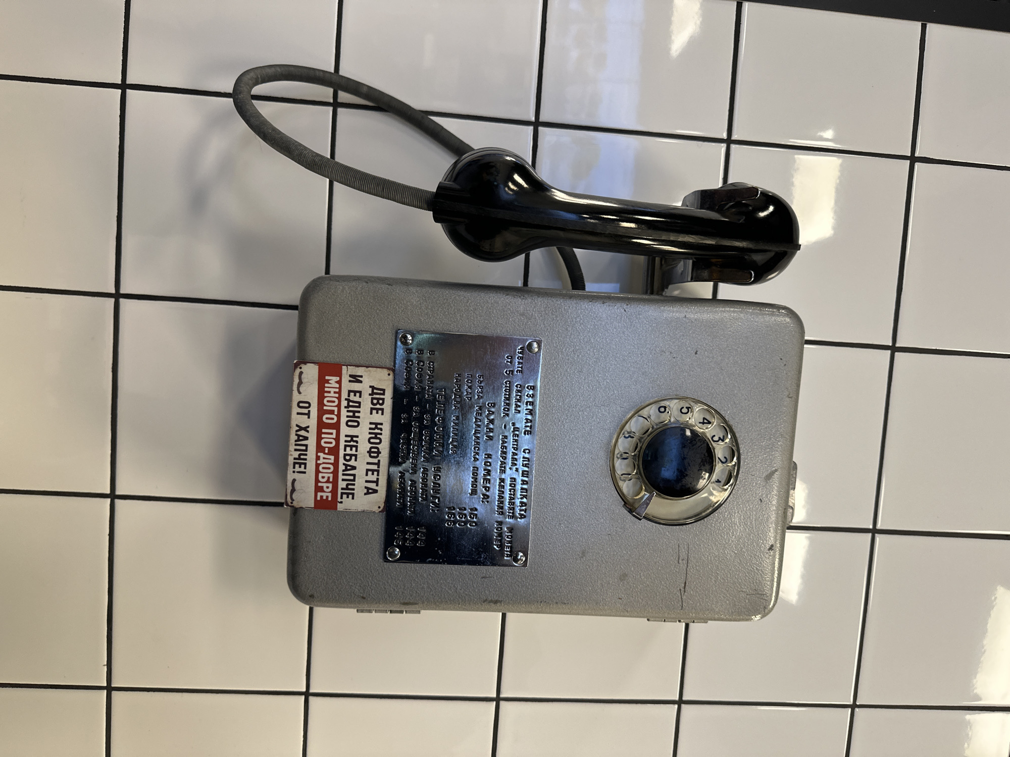 The image shows a vintage, coin-operated public telephone mounted on a white tiled wall. The telephone is grey, rectangular, and features a black handset connected by a grey coiled cord. A rotary dial is visible on the front of the phone. Two metal plates are attached to the front of the phone. One contains instructions and emergency numbers in Bulgarian. The second contains an advertisement in Bulgarian. The wall is covered in white tiles with black grout lines.
Based on the Bulgarian language and the emergency numbers listed, the telephone is located in Bulgaria, likely the city of Sofia.