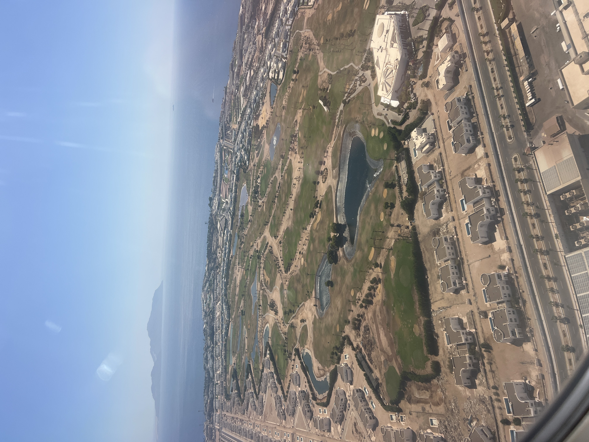 This aerial photograph captures a section of Sharm El Sheikh, Egypt, showcasing a coastal resort area characterized by extensive golf courses and residential developments. In the foreground, a row of modern white villas with flat roofs and individual swimming pools are arranged along a paved road. Beyond the villas, a sprawling golf course dominates the landscape, featuring manicured green fairways, sand traps, and several artificial lakes. The course is interspersed with scattered palm trees and arid, undeveloped land. Further in the distance, the coastal city unfolds with a dense cluster of hotels and other resort buildings, many featuring white architecture. The deep blue of the Red Sea forms the backdrop, meeting the horizon under a clear, bright blue sky. Islands are visible in the far distance, and a small ship is seen on the water. The overall impression is one of a well-developed tourist destination in a desert coastal environment.
