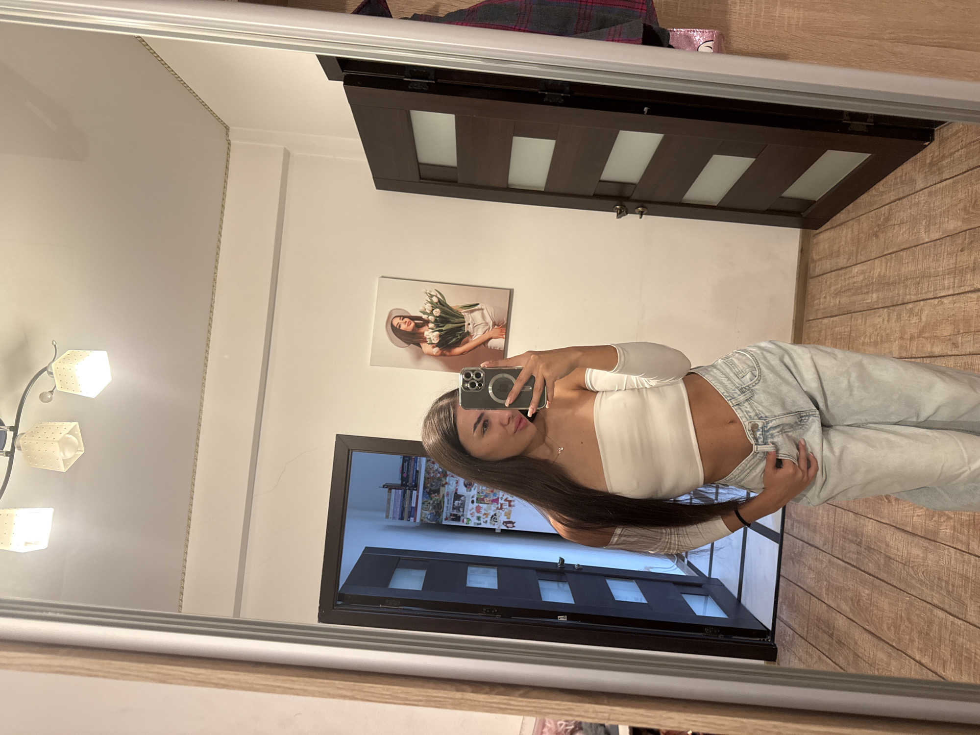 The image is a full-length mirror selfie taken indoors, likely in a residential setting. A young woman with long dark hair is standing in front of the mirror, wearing a white off-the-shoulder crop top and light-wash baggy jeans. She is holding a smartphone in her right hand, taking the photo. Her left hand is in her jeans pocket. In the reflection, a door with frosted glass panels is visible to the left, and another dark wood door with similar frosted panels is to the right. A piece of artwork depicting a woman holding a bouquet of tulips hangs on the wall between the doors. The room has wooden flooring and a ceiling light fixture with three square shades. The overall impression is of a casual, personal snapshot. The location is not identifiable from the image itself, and therefore cannot be specified as Chișinău, Moldova.