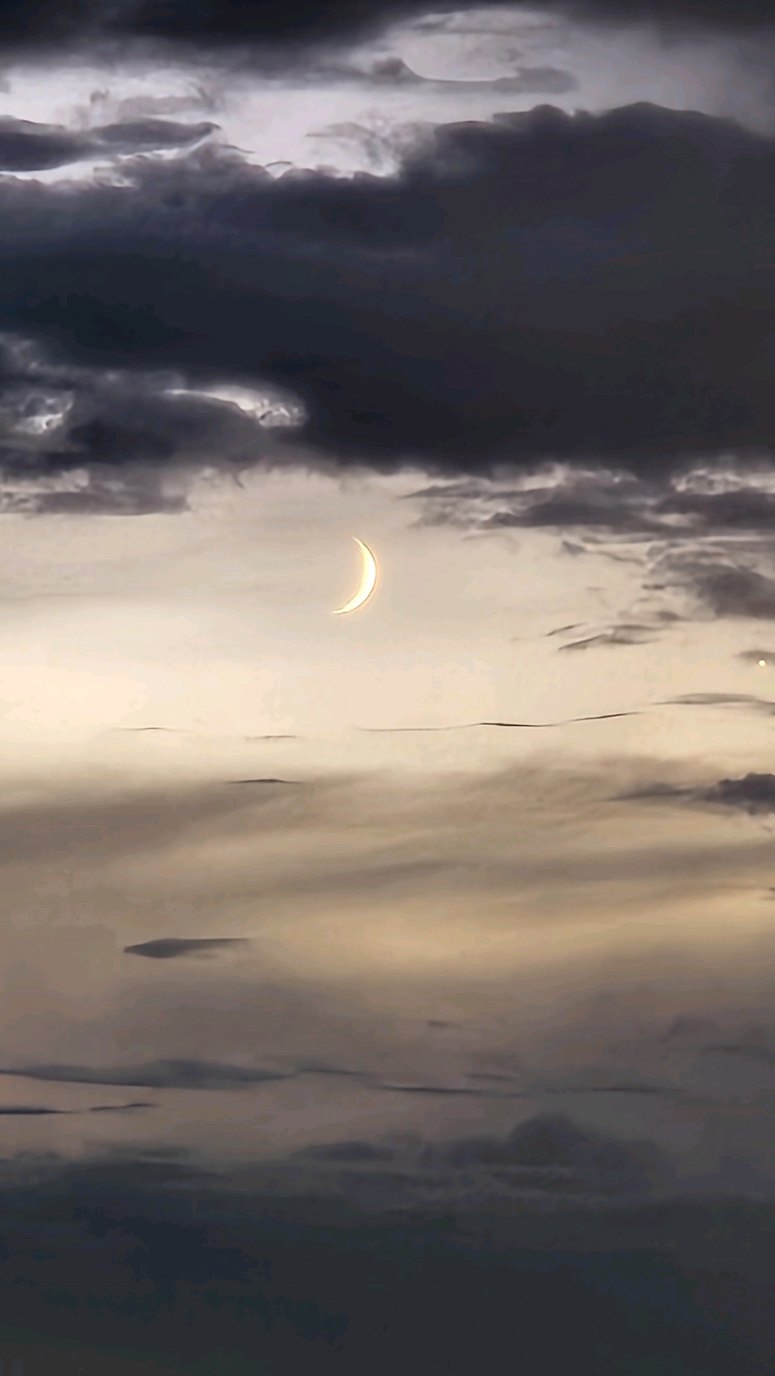 The image depicts a crescent moon in a cloudy sky above what appears to be the coast of Općina Tučepi, Croatia. The moon, a bright sliver of light, is positioned in the upper center of the frame, surrounded by varying shades of gray and white clouds. The lower portion of the image shows darker, more ominous clouds, suggesting a transition in weather or time of day. There are no people, objects, or discernible structures visible in the scene. The overall impression is one of a serene, natural landscape at dusk or dawn, with the celestial body being the focal point against a dramatic sky.