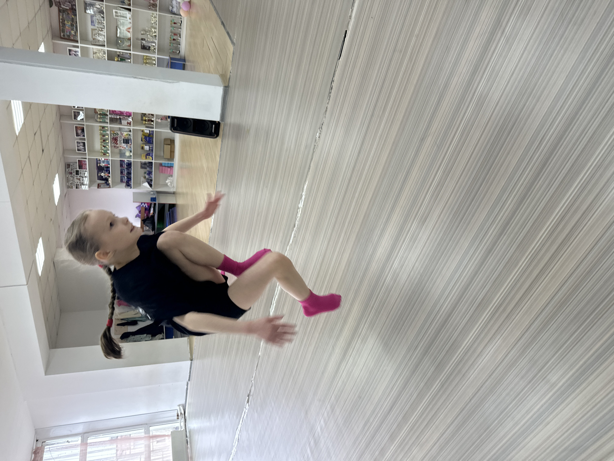 A young girl is captured mid-action indoors, likely performing a gymnastic, dance, or acrobatic move. She has light brown hair styled in two braids, wears a black short-sleeved top, black shorts, and bright pink socks. Her body is angled, with one leg bent and raised, and her arms are outstretched, conveying dynamic movement.
The setting appears to be a training or practice facility. The floor is light-colored, featuring distinct parallel striped patterns, possibly a specialized sports surface. In the background, there is a large white shelving unit filled with numerous trophies, awards, and possibly framed pictures, indicating a history of achievement at this location. A black speaker is visible on one of the shelves. A window is also visible in the lower left portion of the background, along with a white ceiling featuring recessed lighting panels. The overall impression is that of an active, well-equipped sports or performance studio in a location like Votkinsk, Russia. There is no visible text in the image.