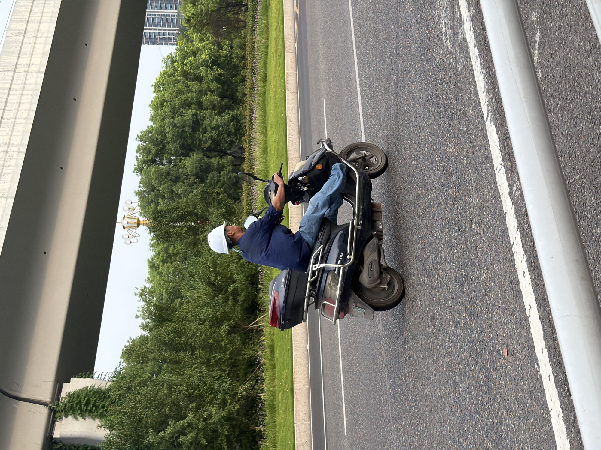 A person wearing a white hard hat and blue shirt rides a dark-colored scooter on a paved road. The scooter has a black storage box on the rear. The person is wearing blue jeans and a blue shirt. The scene takes place outdoors on a road with white lane markings. In the background, there are green trees, a bridge, and buildings. The overall environment suggests a street scene in a city.