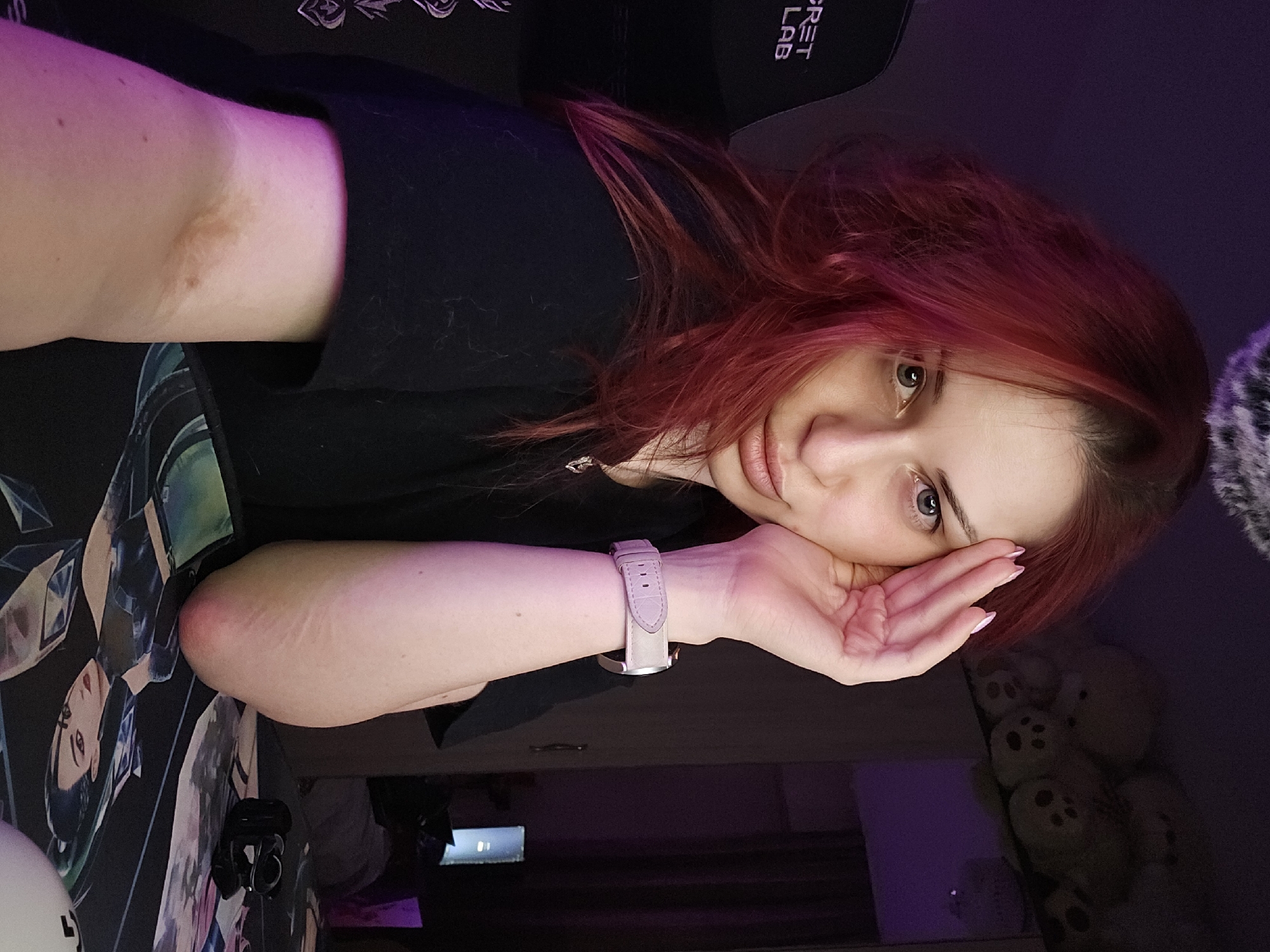 The scene is located in Burgas, Bulgaria. A young adult female with long, reddish-auburn hair and a light complexion is shown in a medium shot, looking directly at the camera. She is wearing a black short-sleeved top and a silver-colored necklace with a small pendant. Her left hand, with nails painted light pink, is positioned near her temple. A watch with a light pink leather strap and a silver-colored casing is visible on her left wrist. On the inner side of her right forearm, a distinct brownish discoloration is present near the elbow, along with several faint linear scars. Her right elbow rests on a dark-colored desk mat featuring a graphic design of a stylized female character. A white object and a small black electronic device are also visible on the desk mat. The subject is seated in a black gaming chair with the "SECRET LAB" logo visible on the headrest. The background is dimly lit with a predominant purple ambient light. A shelf in the upper right background holds several light-colored plush toys. Further back on the right, a reflective surface and a darker room with an illuminated computer monitor are partially visible. A fluffy, dark-colored object hangs from the ceiling in the upper center of the frame.
