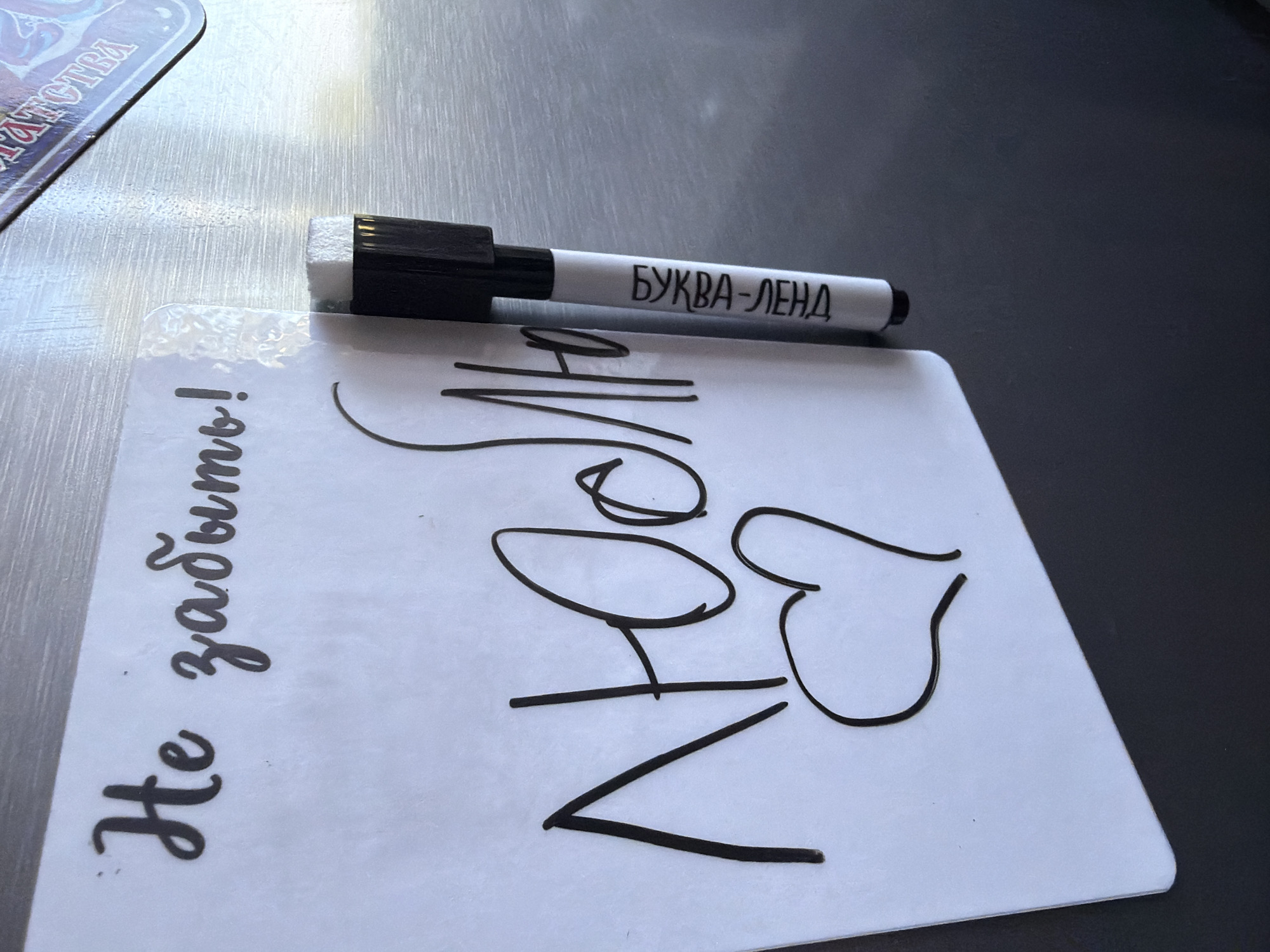 The image depicts a white magnetic board with handwritten text. In the upper left corner, the words "Не забыть!" (Don't forget!) are written in cursive. Below this, the word "Люблю" (I love) is written in larger, bold cursive, with a heart symbol drawn underneath it. A black and white dry-erase marker is attached to the right side of the board. Text on the marker reads "БУКВА-ЛЕHA" (Letter-LEHA). The board is positioned against a textured, grey surface, possibly a refrigerator or a metallic cabinet. The overall scene suggests a personal message or reminder, possibly in a domestic setting. There is no visible indication of the specific location within the city of Chaikovsky, Russia.