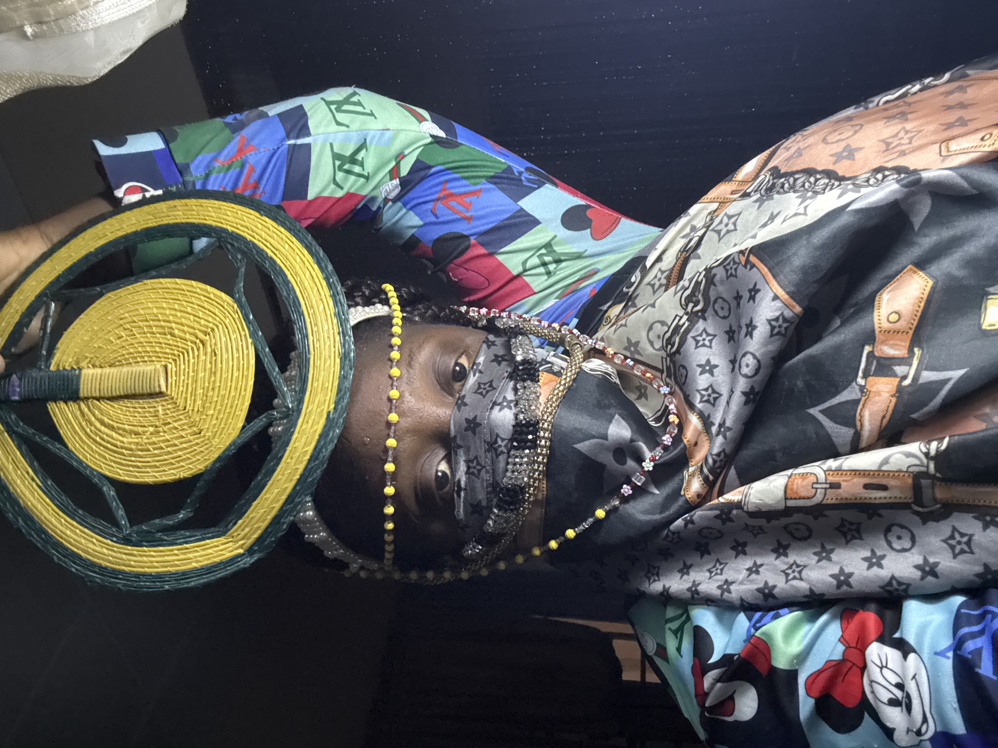 A person is lying down, facing the camera, wearing various layers of patterned clothing and beaded jewelry. The clothing includes items with Louis Vuitton logos, Mickey Mouse prints, and belt buckle designs. The person has a yellow and green coiled circular object near their head. The background is dark and blurry, obscuring details of the environment. No specific location within the city of Madangala, Nigeria, is discernible from the image.