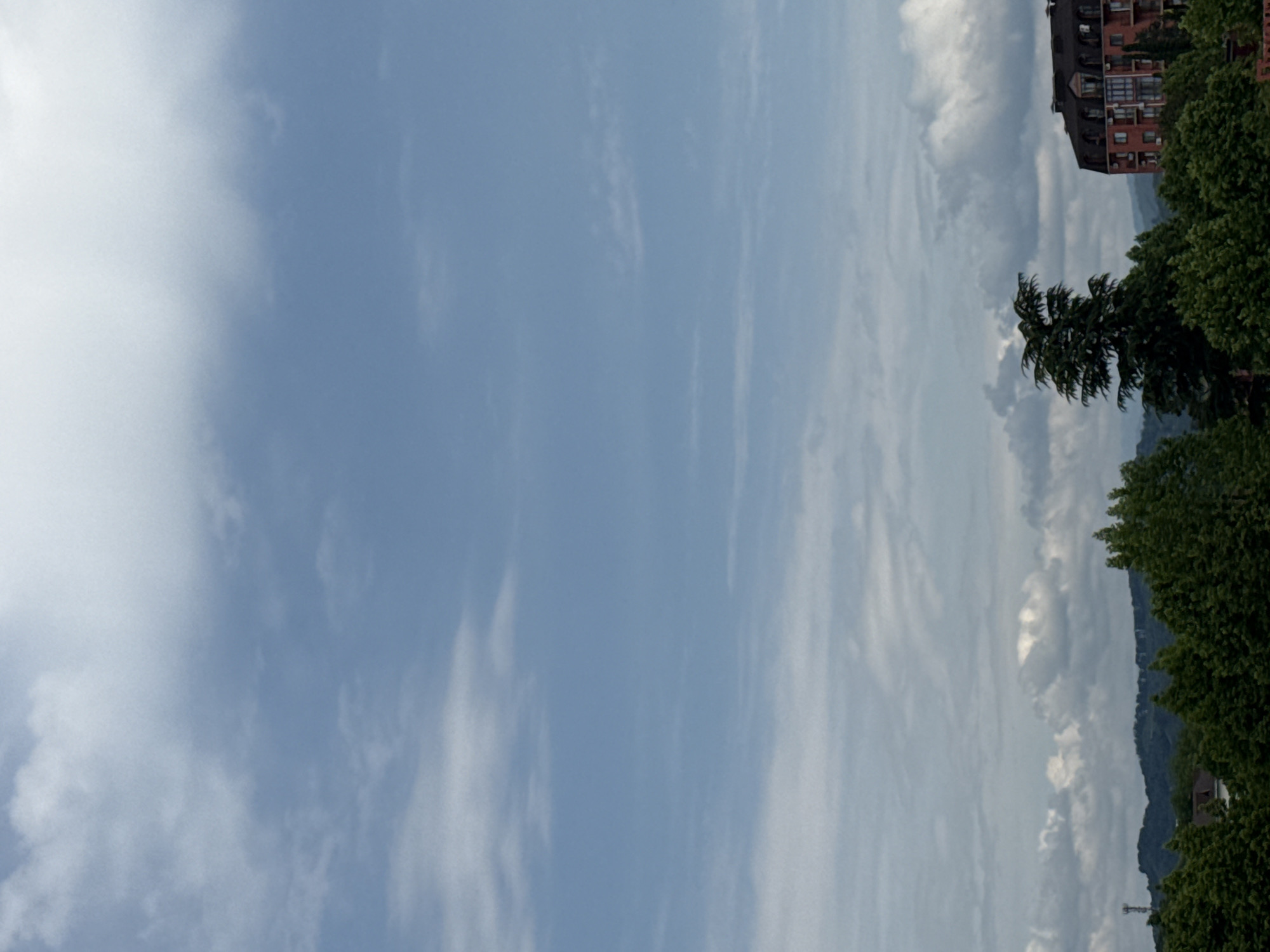 The image captures a view of a cityscape in Sochi, Russia, dominated by a vast, cloudy sky. In the foreground, lush green trees form a dense canopy, obscuring much of the lower urban landscape. A single, dark-roofed brick building with multiple windows is visible peeking through the trees on the right side of the frame. In the distance, rolling hills are visible, also covered in trees. The sky is a mix of blue and white, with various cloud formations, including wispy cirrus clouds and heavier cumulus clouds. There are no people visible in the image.
