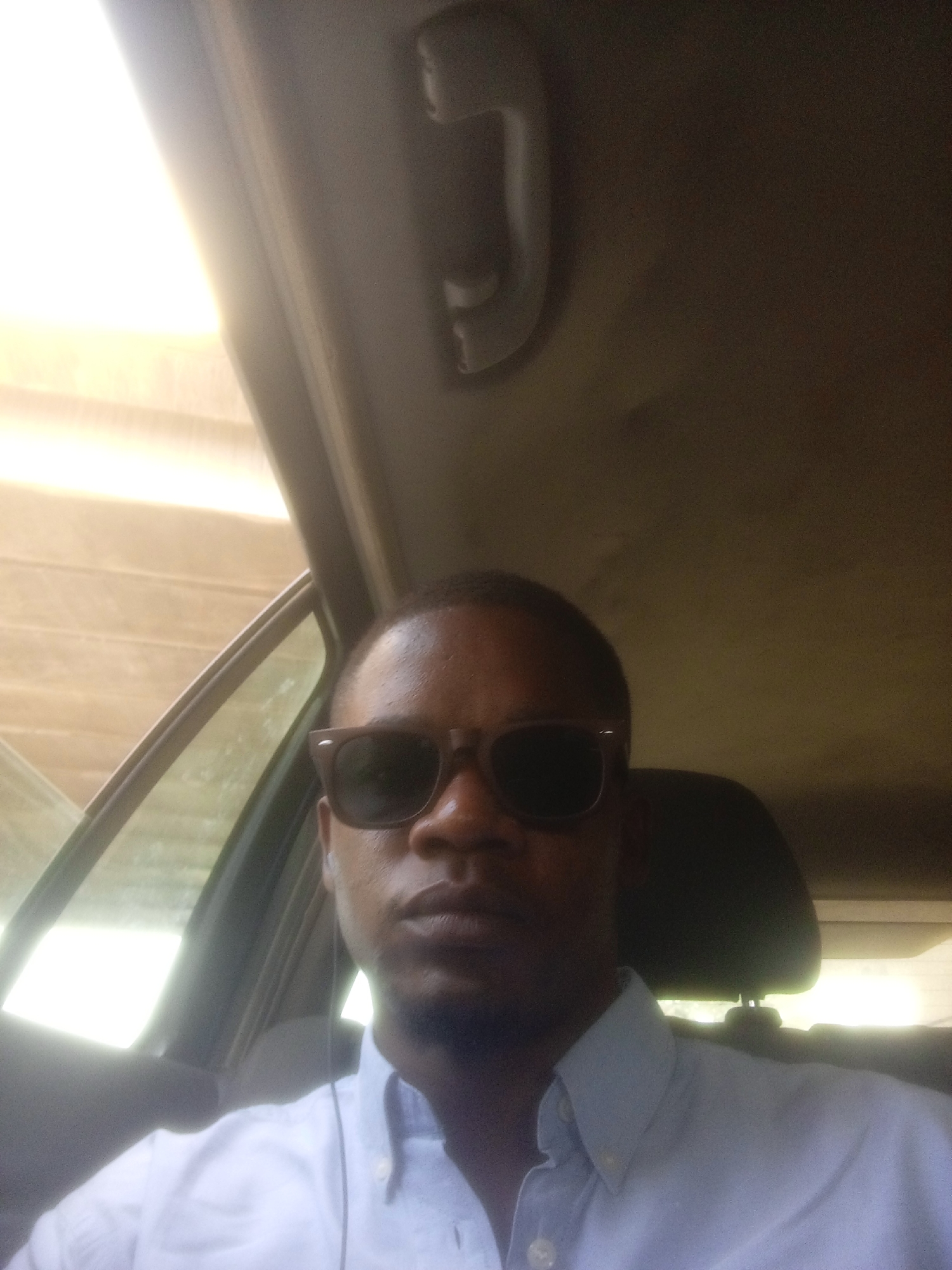 A medium, close-up shot depicts a Black male inside of a car. He is wearing sunglasses and a button-down shirt. The car door and window are visible to the left, and a headrest is visible behind him. The image does not provide the location in the city of Unknown, Unknown.