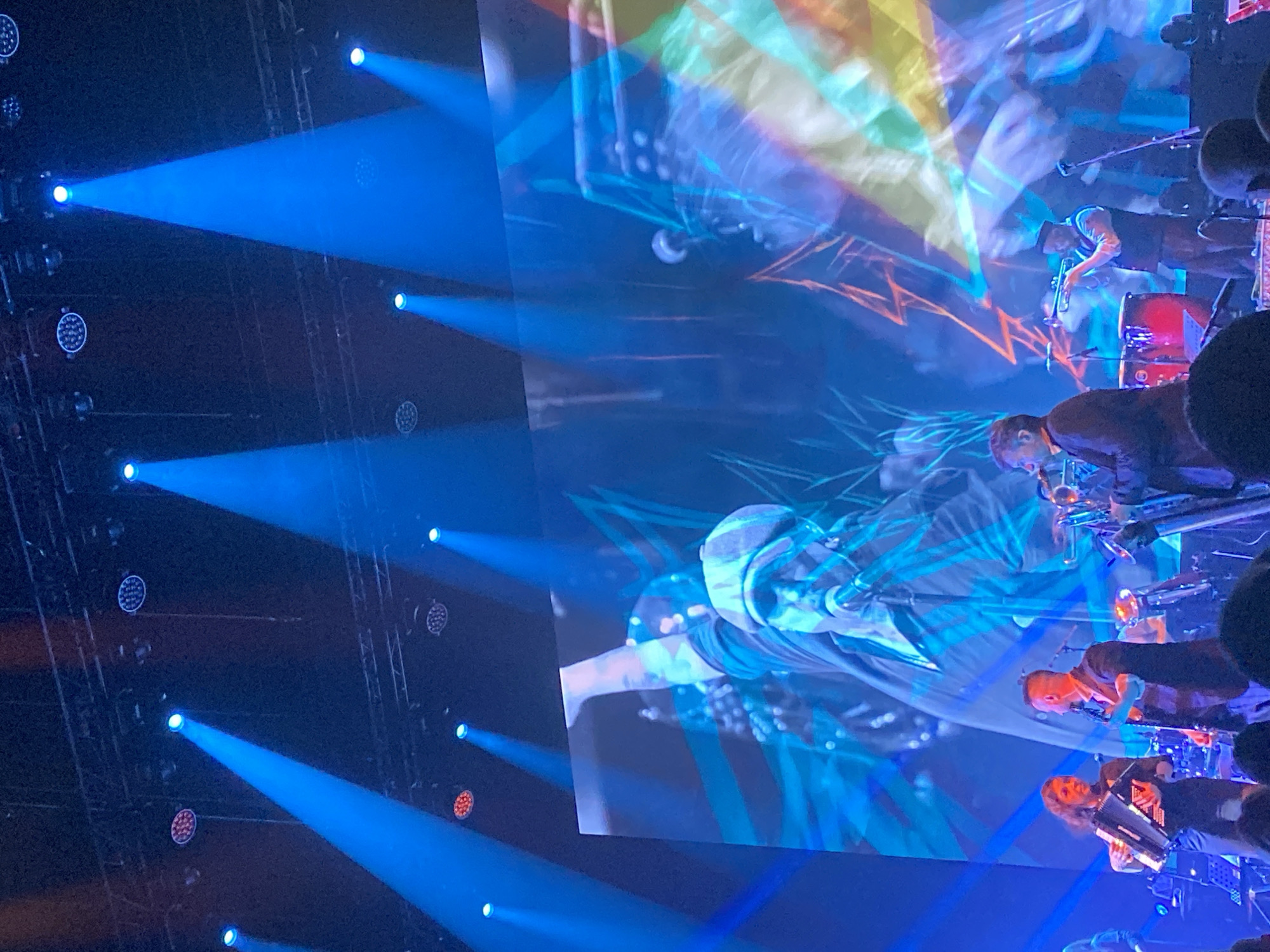 The image depicts a concert scene, likely at the Yekaterinburg Expo International Exhibition Center in Yekaterinburg, Russia, judging by the presence of a large stage, lighting rigs, and a screen projecting a band performance. Several musicians are visible on stage, playing instruments such as trumpets, saxophones, and accordions. The stage is illuminated by numerous blue spotlights. The screen displays a live or prerecorded performance of the same band. The foreground shows partial silhouettes of audience members. The overall atmosphere suggests a live musical performance.