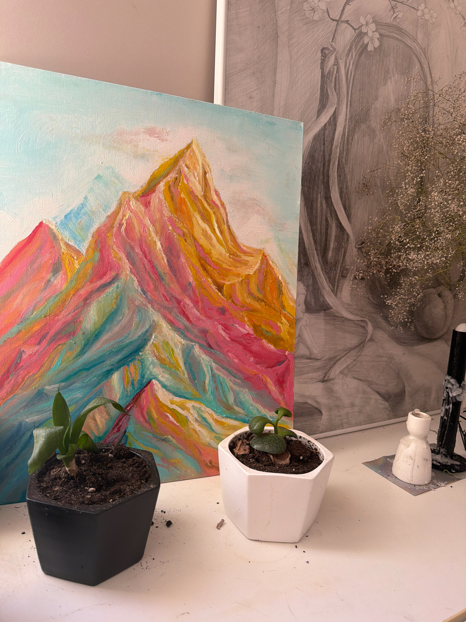 The image depicts a tabletop arrangement with two small potted plants in the foreground. To the left is a black hexagonal pot containing a small green plant with two leaves. To the right is a white hexagonal pot with a similar small green plant. Behind the plants, a colorful painting of mountains in shades of pink, orange, blue, and yellow is leaned against a larger, grey pencil sketch of a figure draped in fabric, with a branch of delicate white flowers above. To the far right, a black candle holder with a partially burned candle and a small white ceramic figurine are visible. The surfaces appear to be clean and the lighting is soft. There is no indication of the city of Samara, Russia in the image.