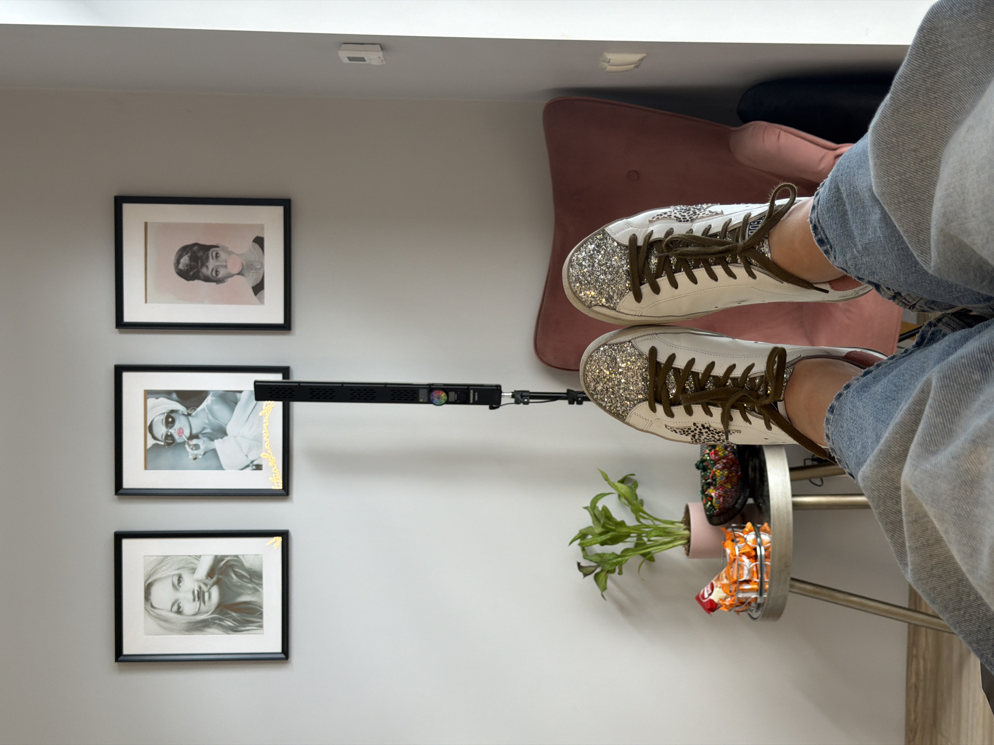 The image shows a person sitting with their feet on a round side table, inside a room with gray walls. The person is wearing jeans and glittery white sneakers with brown laces. Behind them is a pink chair and a black object is visible near the top. On the wall are three framed pictures of women. On the side table, there is a potted plant, a black object decorated with colorful beads, and a bag of Cheetos. A long black light is positioned above the table, pointing toward the wall with the pictures. The image was taken indoors, in Riga, Latvia.