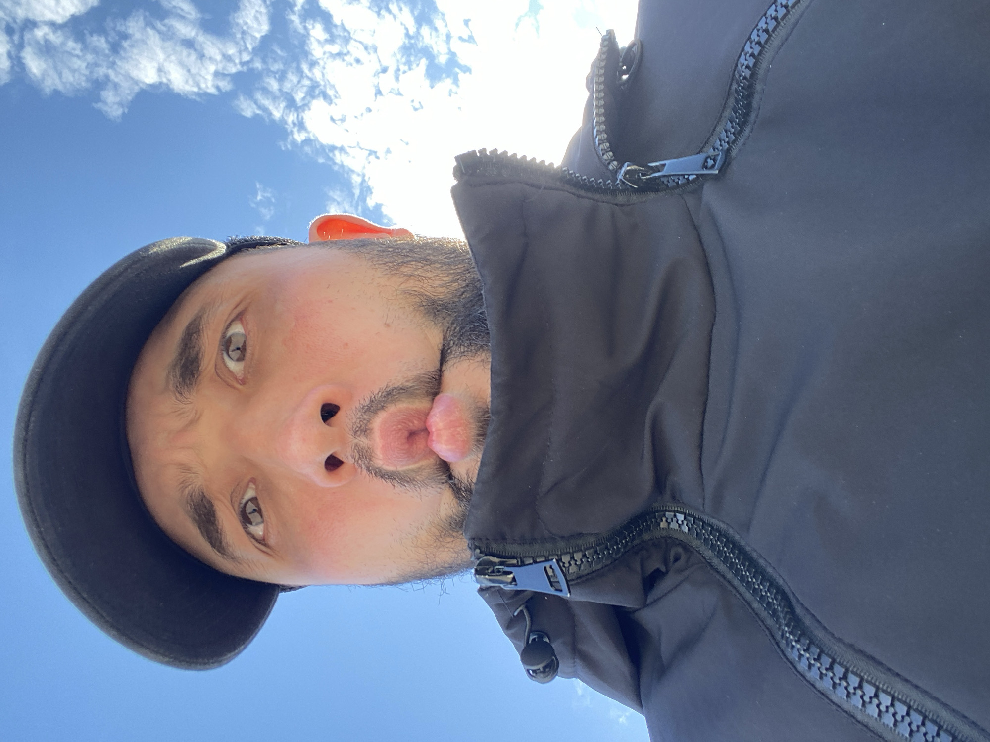 A man with a beard and mustache, wearing a black cap and a black jacket with a zipper, is looking up at the camera with a puckered mouth expression. The background is a blue sky with white clouds. The photo appears to have been taken from a low angle. There is no information in the picture that can confirm its location to be Koskolovo, Russia.