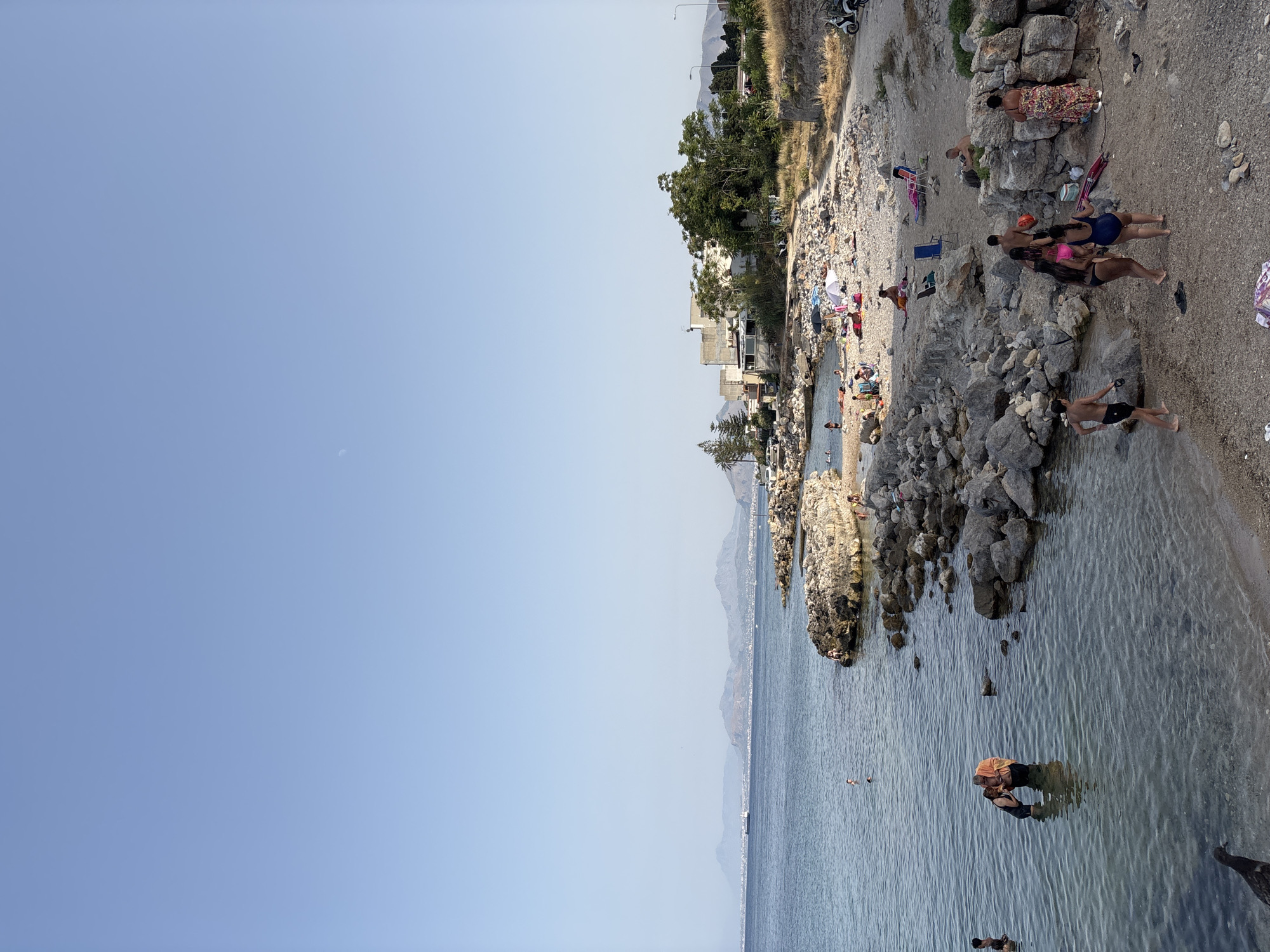 The image depicts a rocky beach with people enjoying the water and the shore. In the foreground, the clear, shallow water laps against the sandy and rocky coastline. Several individuals are visible wading in the water or standing on the beach. One couple is interacting in the shallow water, with one person wearing an orange garment. Further along the shore, a group of young people are gathered, with some in swimwear.
Beyond the beach, a rocky embankment leads up to a residential area with buildings and vegetation. In the distance, a hazy mountain range is visible, suggesting the presence of a larger city or landscape beyond the immediate coastal scene. The sky is clear and pale blue, with a faint crescent moon visible. The overall impression is a relaxed, summertime scene at a coastal location.
While the image itself doesn't explicitly state its location, the architectural style of the buildings and the landscape are consistent with coastal areas around Palermo, Italy. The rocky coastline and the Mediterranean Sea are characteristic of the region. The presence of a ship on the horizon could indicate a port or busy shipping lane, which is also common in the vicinity of Palermo.