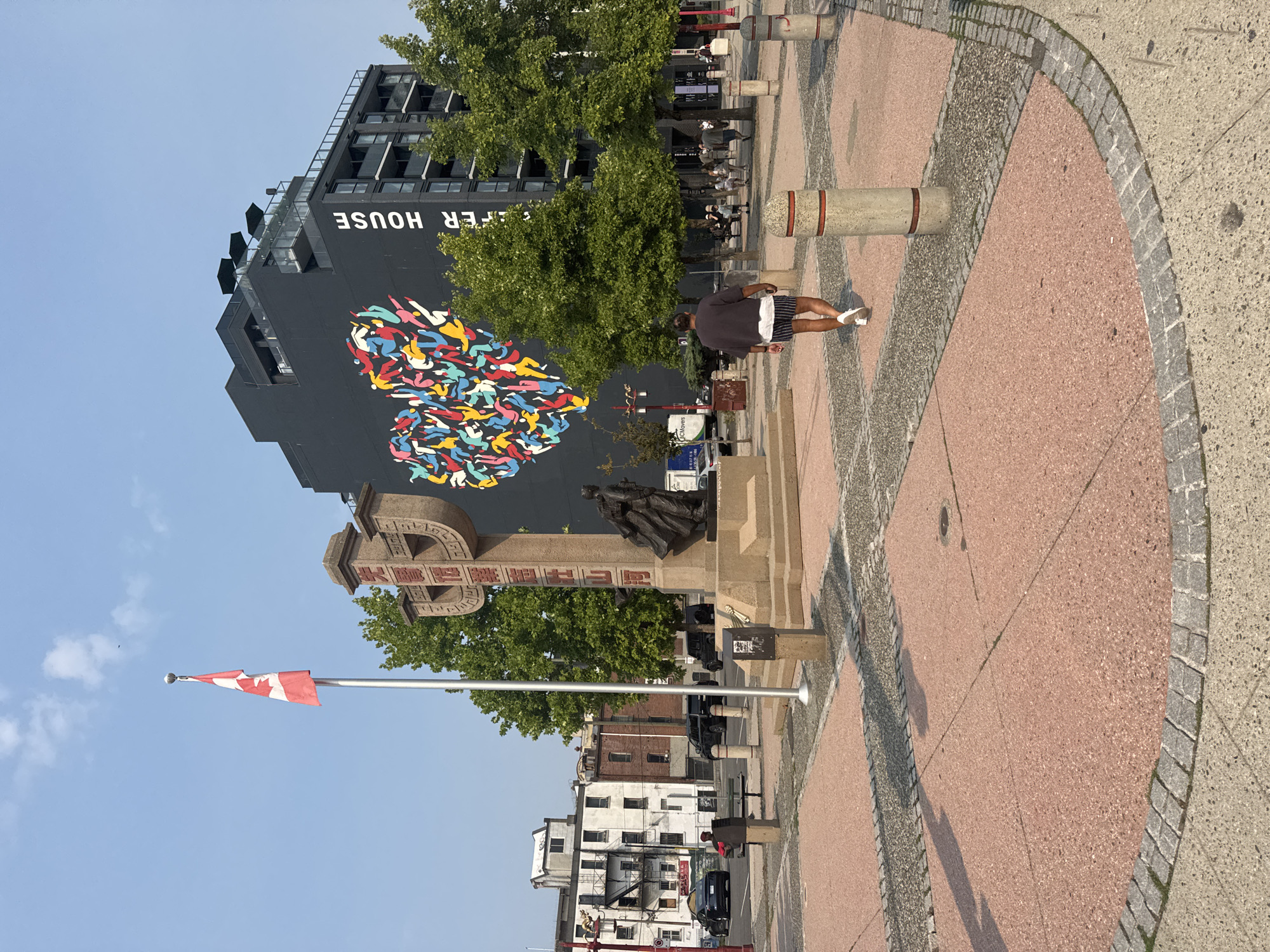 This image depicts a sunlit public square with a statue and a large mural on a building in Vancouver, Canada. In the foreground, a man walks away from the camera, wearing a dark t-shirt, striped shorts, and white sneakers. The square is paved with pinkish aggregate and bordered by cobblestones. A flagpole with a Canadian flag stands to the left of a bronze statue on a stone pedestal. The statue appears to depict a figure in a cloak. Behind the statue, a dark, multi-story building is prominently featured, adorned with a large, colorful mural forming a heart shape composed of abstract human figures. Text reading "FER HOUSE" is visible on the building's facade. Trees line the area, and in the background, older buildings and parked vehicles are visible, suggesting an urban setting. The sky is clear and blue.