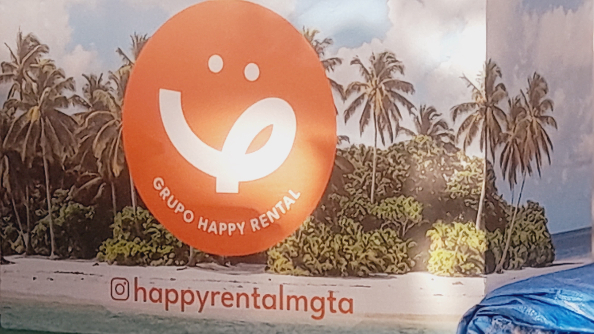 This image displays a vibrant advertisement or branding material featuring a tropical beach scene. At the center, a large, bright orange circular logo immediately draws the eye. The logo contains a stylized white smiling face, which also abstractly forms a shape resembling a lowercase "q" or an abstract swirl, with two small white dots representing eyes. Curved around the lower half of the orange circle, the white text "GRUPO HAPPY RENTAL" is clearly visible, indicating the name of the business.
The background of the advertisement depicts a picturesque, sunny tropical landscape. Numerous tall palm trees with dark trunks and fronds dominate the midground, interspersed with lush green foliage and shrubs. Above the trees, a light blue sky with soft white clouds suggests pleasant daytime weather. The bottom of the scene shows a sandy beach area stretching across, with calm, light blue water visible towards the right edge. This idyllic setting evokes feelings of relaxation and vacation.
At the very bottom of the image, below the tropical scene and the main logo, an Instagram handle is prominently displayed in orange text: "@happyrentalmgt". This handle suggests a social media presence for the company. A small, blurry section of blue material or fabric is partially visible in the bottom right corner of the overall image. The "mgta" in the Instagram handle likely refers to Margarita Island, consistent with the tropical setting in Pedro González, Venezuela.