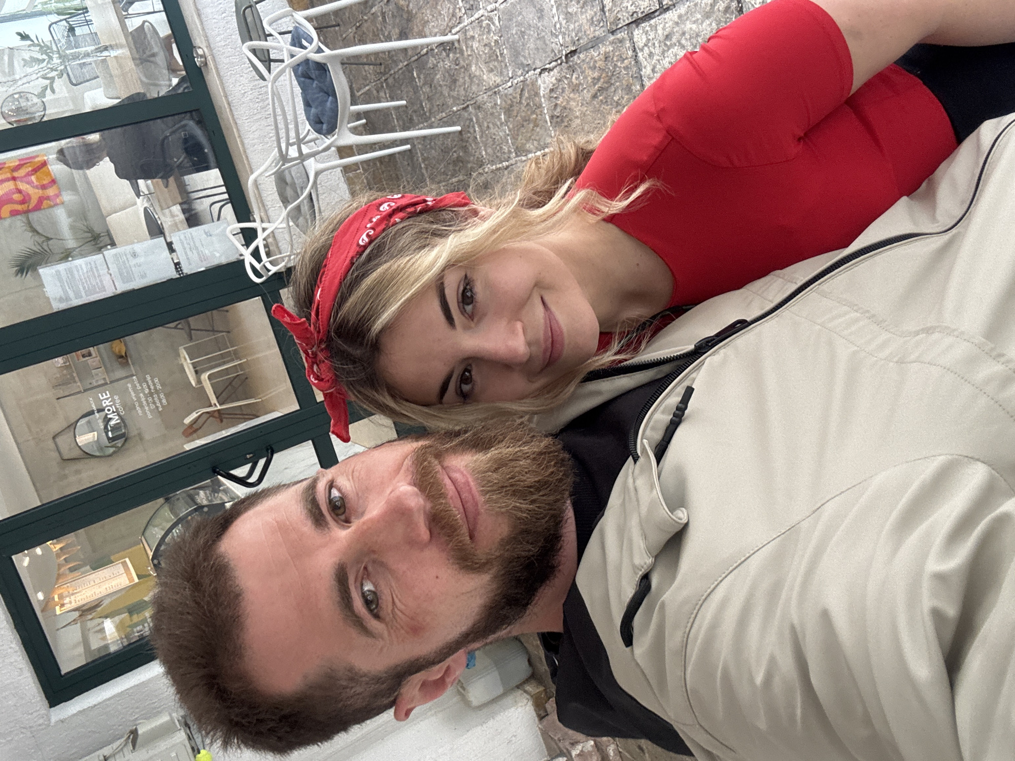 A selfie captures a man and woman posing together outside in Budva, Montenegro. The man, with a beard and wearing a jacket, is closer to the camera. The woman is behind him, wearing a red top and a bandana in her hair. A storefront with windows is visible in the background, as well as chairs and an outdoor seating area, with an advertisement for coffee on a window. The ground is paved with stone.