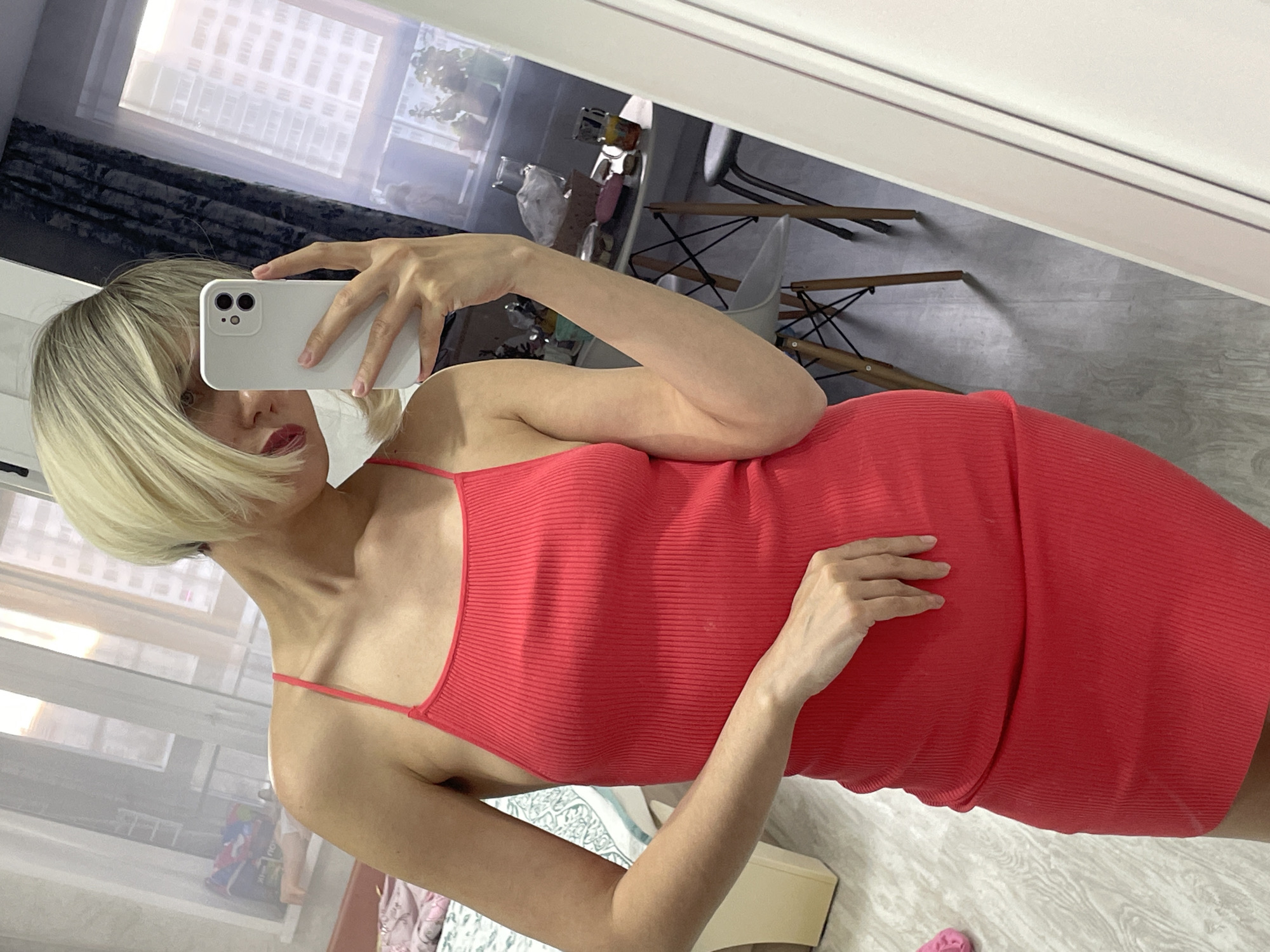 A young person with short, blonde hair holds a white cell phone in front of their face, partially obscuring it. They are wearing a red, ribbed tank dress with thin straps. The location appears to be an apartment interior. Behind the person, a window shows what seems to be a high-rise building, potentially suggesting an urban environment. Based on the available information, it's impossible to confirm that this image was taken in Izhevsk, Russia, since there are no identifying landmarks or metadata. The details include furniture like chairs with wooden legs and a white, rectangular object near the bottom of the frame. A partial view of what could be a bed or other soft furniture is visible in the lower left. The overall impression is that of a selfie taken in a domestic setting.
